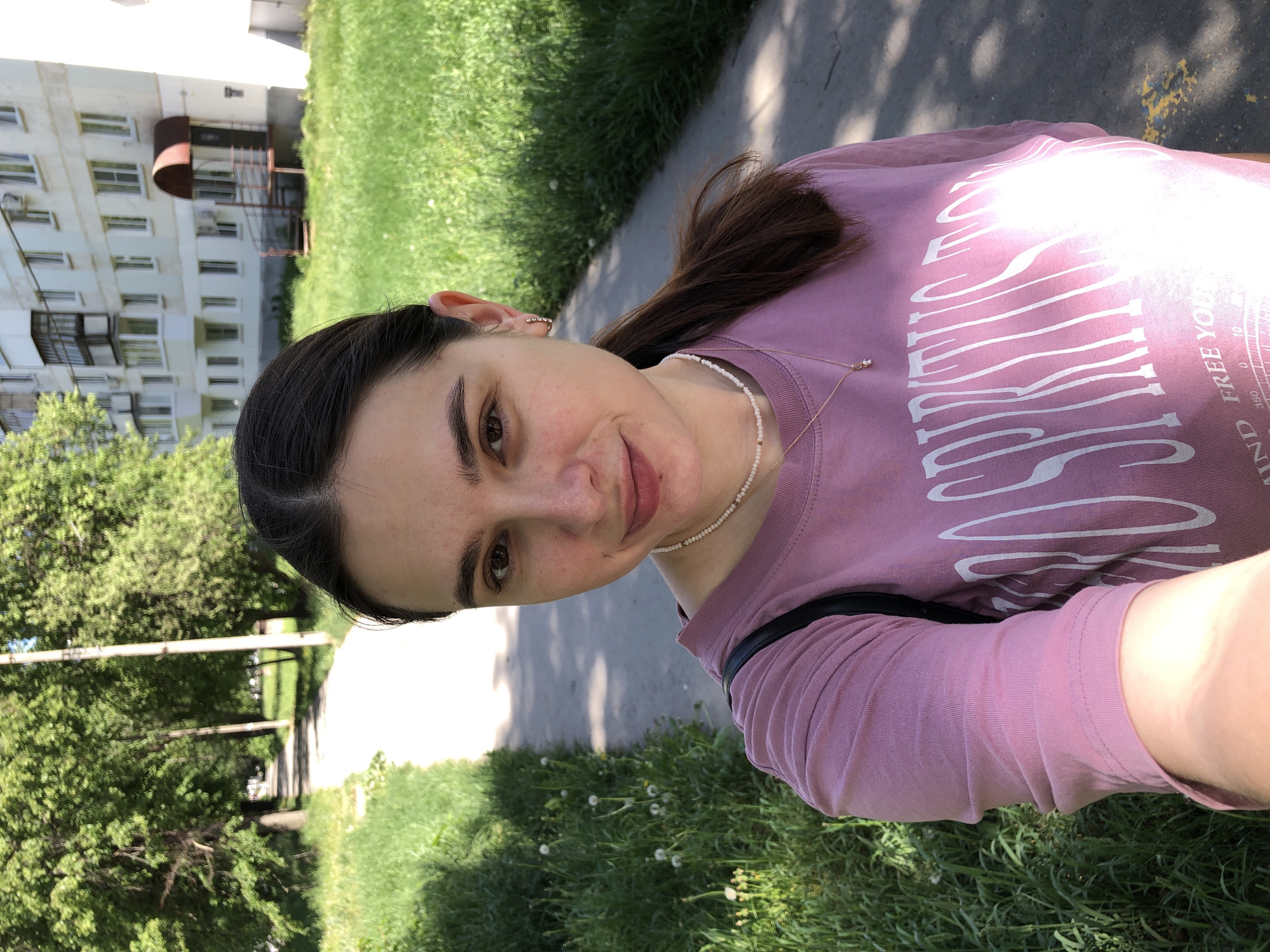 The image is a self-portrait of a woman with fair skin, brown hair pulled back from her face, and brown eyes. She is wearing a pink t-shirt with white graphic text, a white beaded necklace, and a gold pendant necklace. She is standing outside on a concrete path next to a grassy area. In the background, there is a multi-story residential building. The presence of the specific architecture of the building and the foliage, combined with the original prompt information, indicate the location is likely within the city of Chaikovsky, Russia.