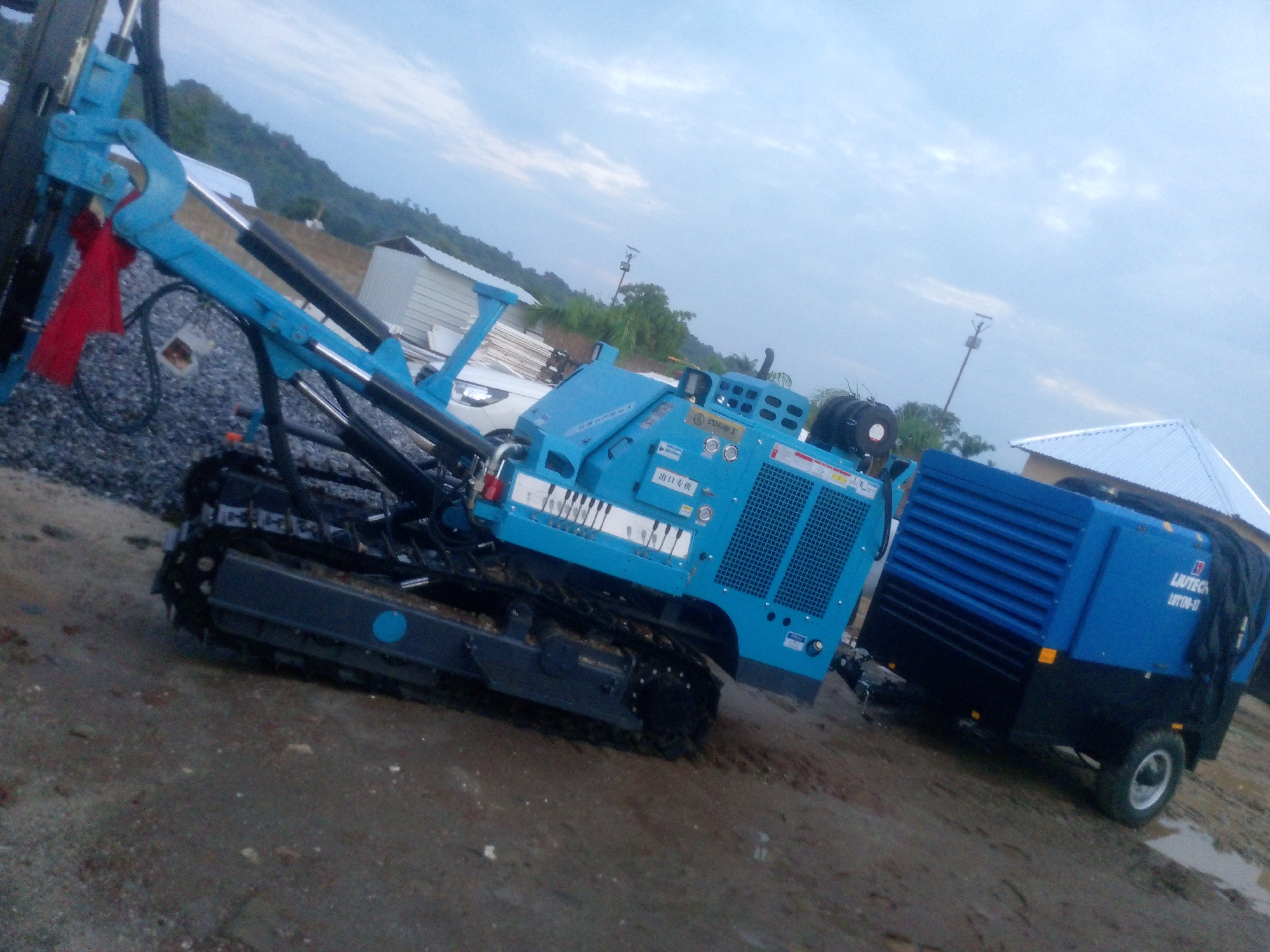 The image captures an outdoor scene in Indo, Nigeria, featuring heavy machinery. In the foreground, a blue drilling rig with a track-based undercarriage is positioned on a dirt and gravel surface. A red cloth is draped over part of the rig's arm. To the right of the drilling rig, a blue and black trailer-mounted compressor unit is visible, with the brand name "LUTECH" and model number "LHT170-HI" printed on its side. In the background, there are buildings with white roofs, lush green vegetation, and a hilly landscape under a cloudy sky. The overall environment suggests an industrial or construction site.