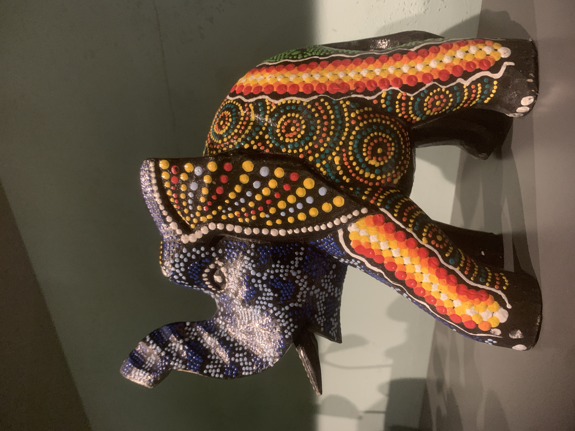 An ornate, multi-colored elephant figurine, likely made of carved wood or a similar material, is prominently displayed against a plain, light greenish-gray wall. The image is oriented sideways, suggesting the object might be wall-mounted or placed on a shelf.
The elephant is intricately decorated with a vibrant dot painting style, reminiscent of aboriginal or folk art. Its body is primarily black, serving as a dark canvas for the colorful patterns. The trunk and ears are adorned with distinct designs. One ear and the lower part of the trunk feature a dense pattern of blue and white dots, creating a textured appearance. The main body and upper parts of the elephant are covered in concentric circles and linear bands of dots in bright colors including red, orange, yellow, green, and light blue. A prominent pattern on the elephant's back consists of horizontal stripes of red, orange, and white dots, interspersed with spiral motifs of yellow, green, and blue dots. White dots are also used to outline certain areas and create additional details.
No activity is taking place; the image captures a static decorative object. The lighting appears artificial, casting soft shadows on the wall behind the figurine. There are no visible people, animals other than the figurine, or specific location cues within the image that would visually confirm the context of Izhevsk, Russia. No text is visible on the object or in the background.