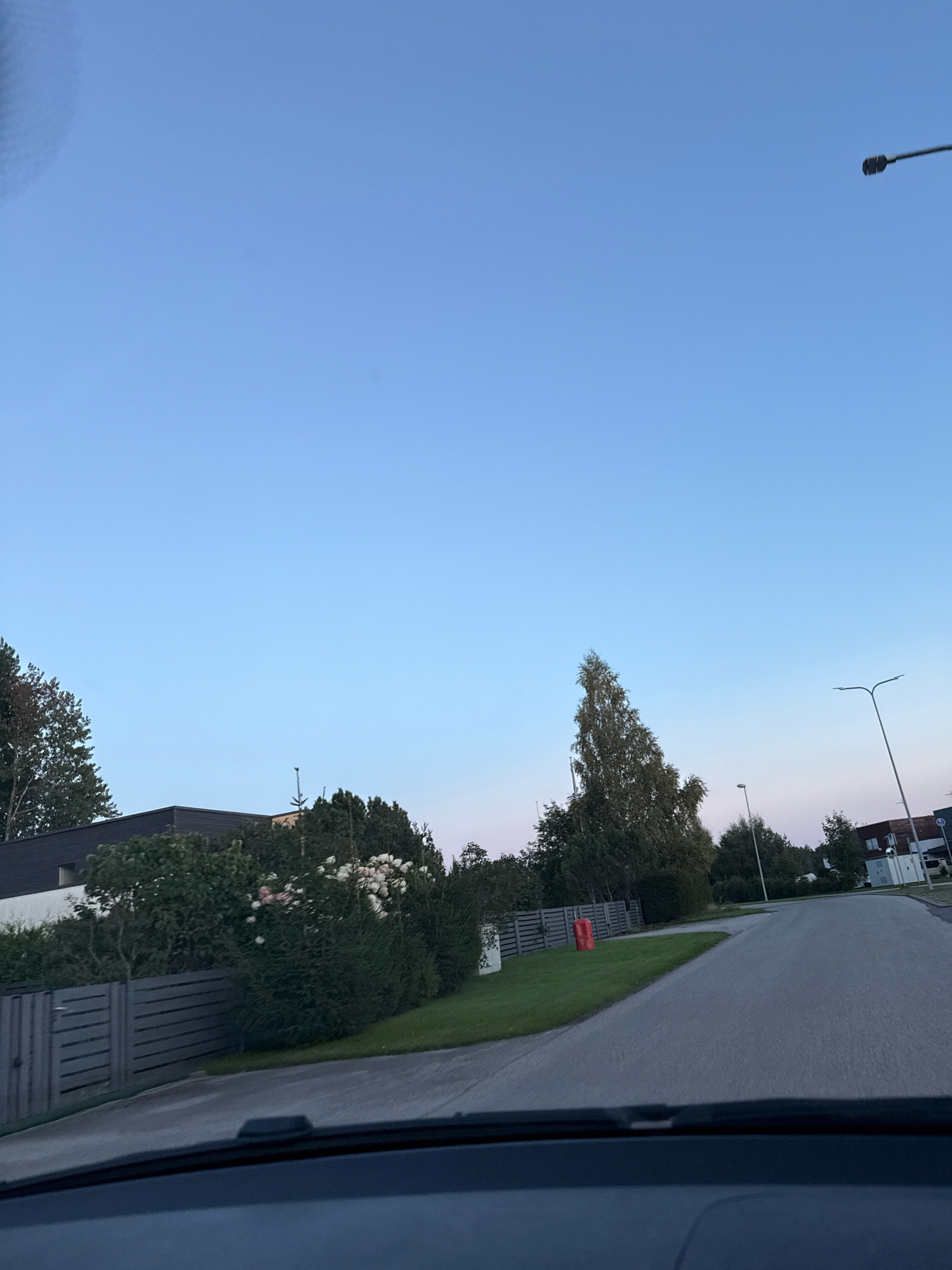 The image shows a street in the city of Alliku, Estonia, likely during the evening or early morning. The sky is a clear, pale blue, transitioning to a lighter hue near the horizon. On the left side of the street, a modern house with a dark facade and white accents is partially visible, surrounded by dense green bushes and trees. A grey wooden fence runs along the property line. A bright red cylindrical object, possibly a mailbox or a decorative element, is placed on the lawn near the fence. Further down the street, more houses and trees are visible, interspersed with streetlights. The road itself is paved and appears to be a residential street. The foreground of the image shows the interior of a vehicle, likely the viewpoint of the photographer, with the dashboard visible.