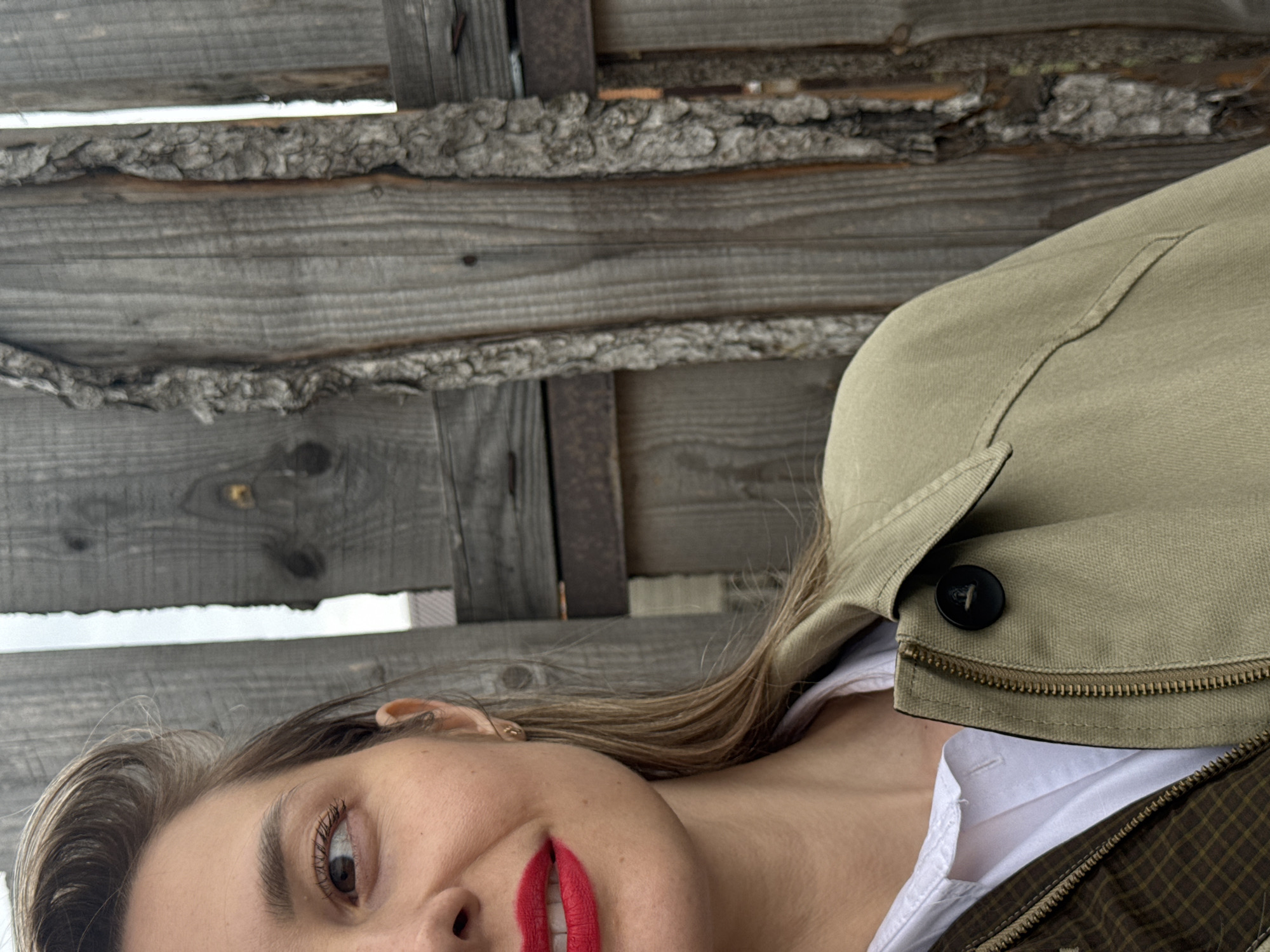 The image depicts a woman with fair skin, brown hair, and red lipstick, positioned in the lower portion of the frame. Her face is angled slightly upwards. She is wearing a white shirt under a light green jacket with a black button. The background consists of a wooden structure made of gray planks and metal elements. There is light visible through gaps in the wood. No specific details indicate the location is Chaikovsky, Russia.