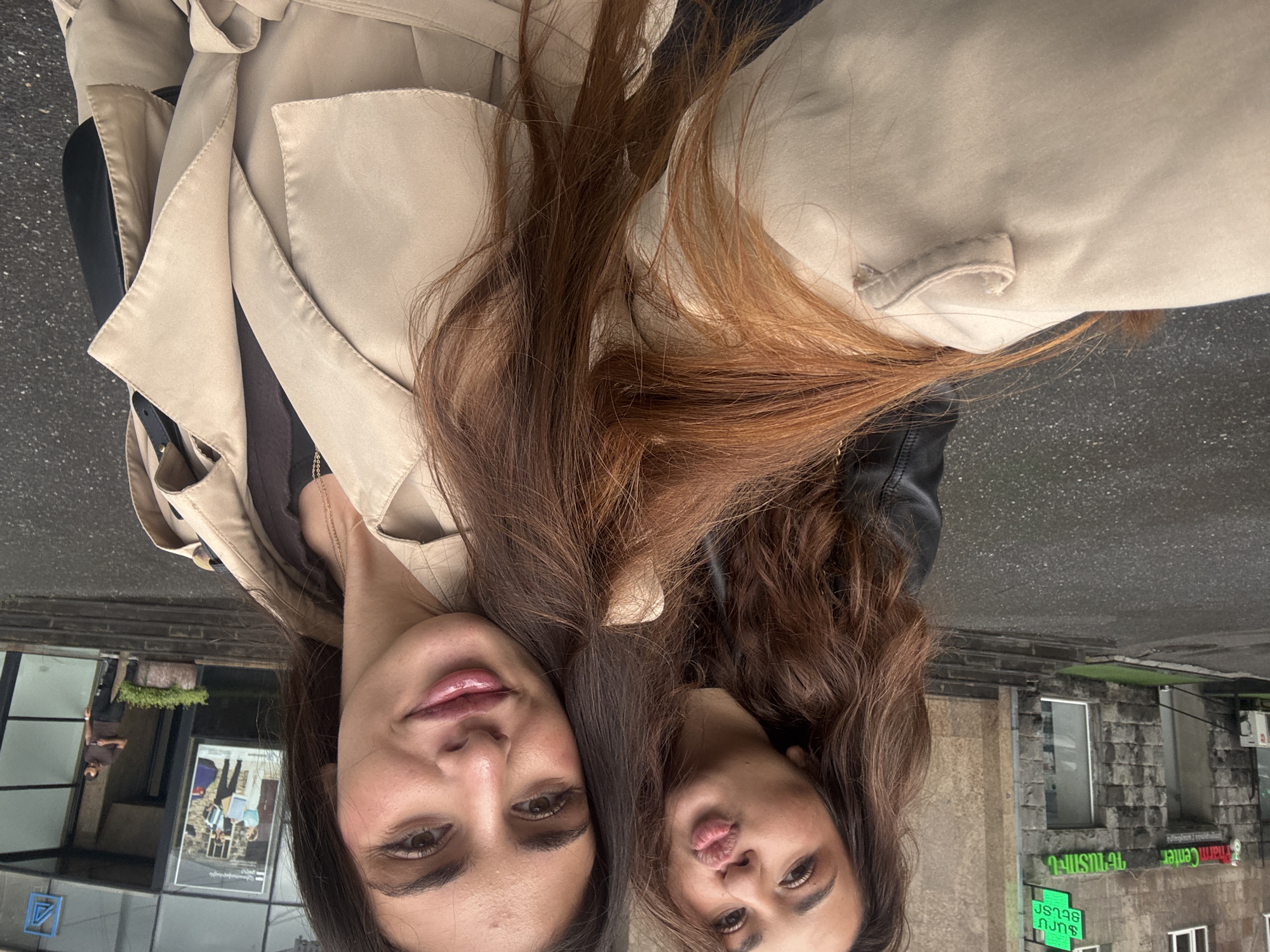 Two women are captured in a close-up, overhead selfie, with the image rotated 90 degrees counter-clockwise. Both are looking down at the camera, their heads close together. The woman on the left (when correctly oriented) has long, dark brown hair, dark eyes, and a pleasant, serene expression with slightly parted, glossy pink lips. She wears a light beige or khaki trench coat over a dark top, with a delicate golden chain necklace visible.
The woman on the right (when correctly oriented) has long, lighter brown or auburn hair, dark eyes, and a playful pouting expression. She is also wearing a similar light beige/khaki trench coat, with what appears to be a black leather jacket collar underneath. Their hair flows around their faces and over their coats.
The background is an urban outdoor setting, featuring street elements and buildings. To the left, reflected in a large glass window, a poster with people is visible, along with a small potted plant and a blue triangular logo. To the right, stone buildings are visible, some with greenery on ledges. The ground or street surface is visible at the top of the frame.
Notable details in the background include visible text on storefront signs, which appear in Armenian script and English. A prominent green sign reads "ԹԱՐՄ ԾԱՂԻԿՆԵՐ" (TARM TSAGHKNER), translating to "Fresh Flowers." Above it, another sign includes the English word "Center" and Armenian script "ՊԱՆԴՈԿ" (PANDOK), meaning "Inn" or "Restaurant." Below the "Fresh Flowers" sign, a partially visible red sign displays "ՇՈՒՇԻԻ" (SHUSHII), likely referring to the city Shushi. The scene is illuminated by natural daylight, suggesting it's daytime. The overall mood is casual and friendly.