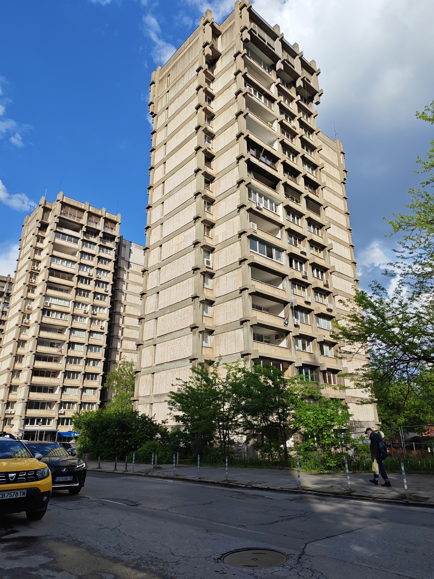 The image depicts a street scene in Sofia, Bulgaria, dominated by brutalist architecture. Two tall, concrete apartment buildings with repetitive horizontal divisions and projecting balconies rise in the background against a blue sky with scattered clouds. A street runs across the foreground, showing a yellow Dacia Duster and a black Lexus SUV parked on the left. A man wearing a dark jacket, jeans, and a face mask walks along the sidewalk on the right, carrying a yellow bag. The sidewalk is bordered by low fencing and vegetation. A manhole cover is visible on the street surface. Vehicle license plates are visible on the cars.