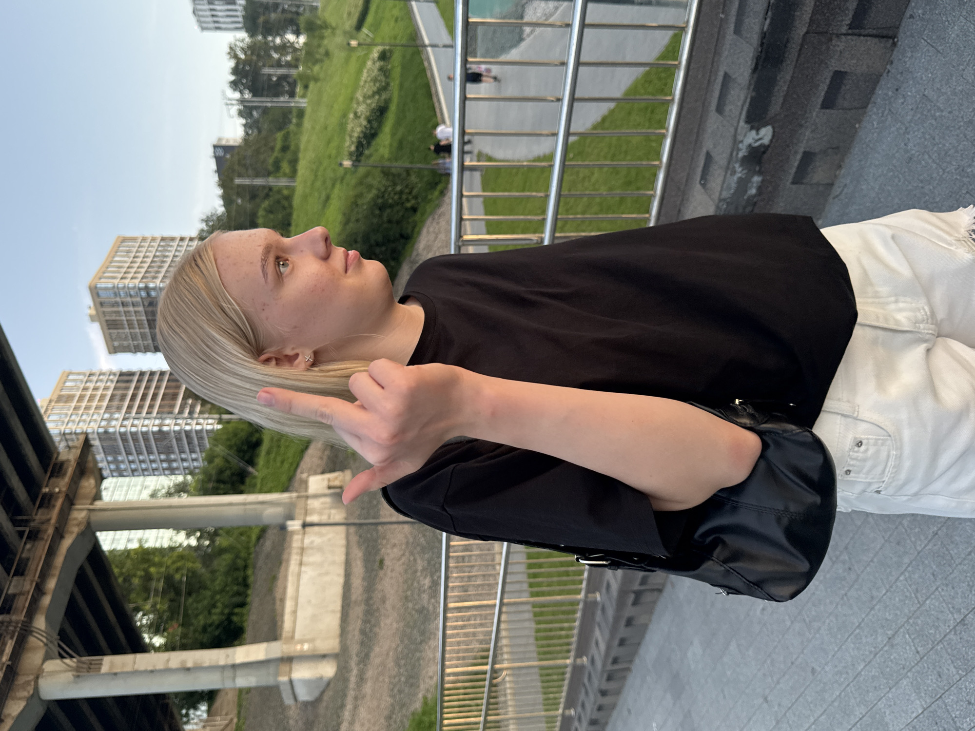 A young woman with blonde hair and freckles stands on a paved walkway outdoors, facing to her right. She is wearing a black t-shirt and white distressed shorts, and carrying a black purse on her left shoulder. Her right arm is raised, and she is extending her middle finger. Behind her, a metal railing runs along the walkway, with a grassy slope and more railings beyond. In the background, a large concrete overpass looms, with buildings and trees visible further away. The sky is a pale blue with hints of sunset. The scene appears to be in an urban park setting, possibly with a waterfront or river nearby, given the presence of water in the distance. The lighting suggests it is late afternoon or early evening. The location is the Embankment of the Kama River in Perm, Russia.