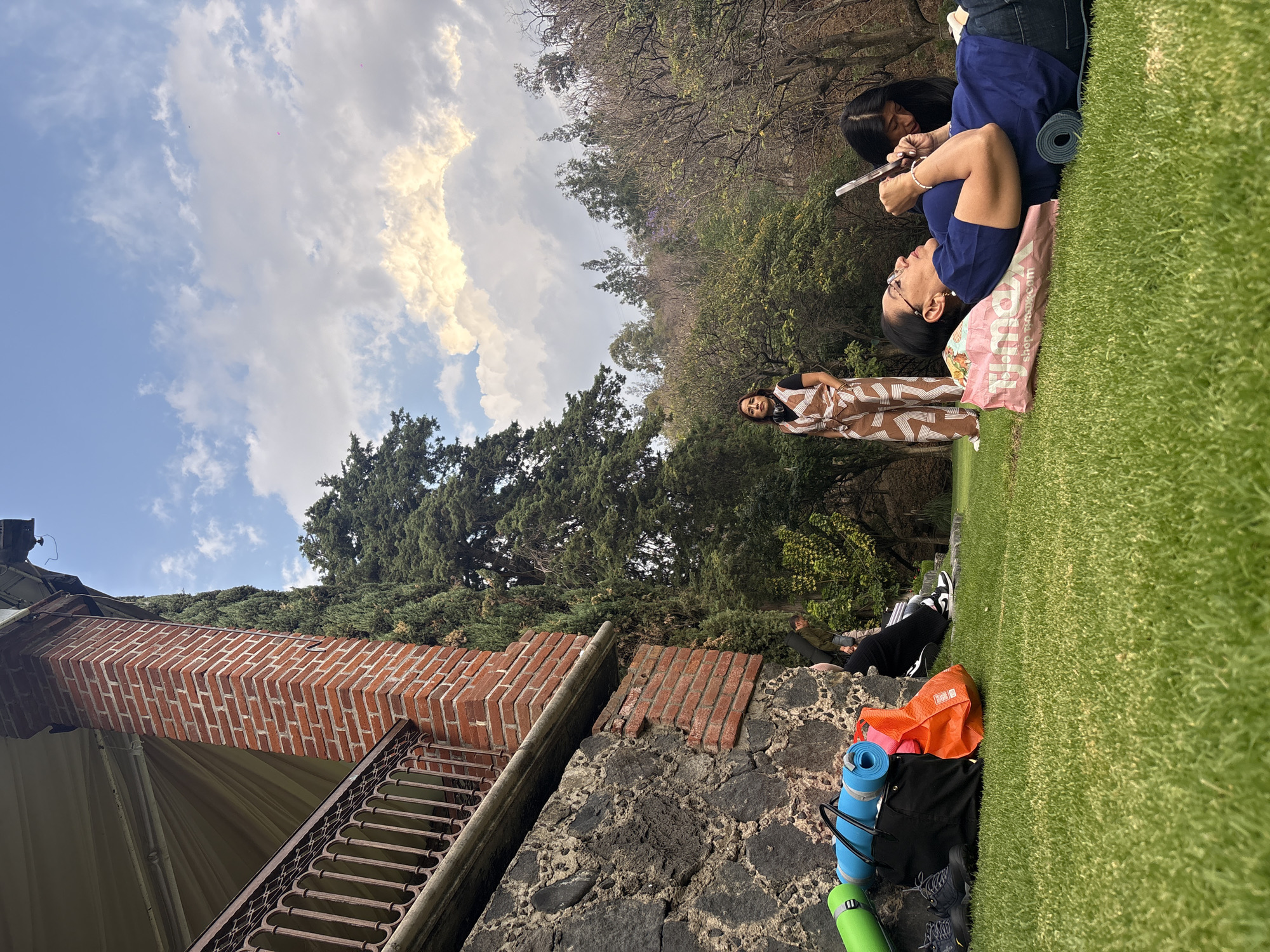 The image depicts a group of people gathered outdoors in Santa Ana Acozautla, Mexico. Two women are lying on the grass, one holding a phone while the other looks on. A woman in a patterned jumpsuit stands nearby. Yoga mats and bags are piled on a stone wall. A building with brick details and a wrought iron railing is visible in the foreground, with trees and a cloudy sky in the background. Other people are visible in the background, sitting on a ledge.