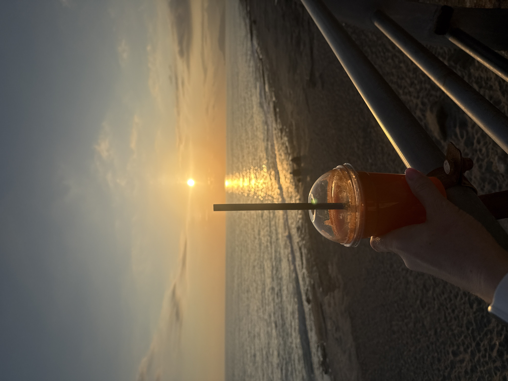 A person's hand holds a drink in a clear plastic cup with a black straw, set against the backdrop of a sunset over the ocean. The image captures a serene moment at what appears to be a beach or shoreline.
The sun, a bright golden orb, is low on the horizon, reflecting a shimmering path of light across the rippling water. The sky above transitions from a warm golden hue near the sun to a softer blue-grey further away, dotted with scattered clouds. On the right side of the frame, dark, diagonal railings or posts suggest the presence of a pier, boardwalk, or similar structure. The sand is visible along the edge of the water.
The hand, adorned with light pink nail polish, holds the orange-colored drink. The dome lid of the cup is clear, allowing the contents to be seen. The overall atmosphere is peaceful and relaxing, indicative of the evening hours. There is no visible text in the image.