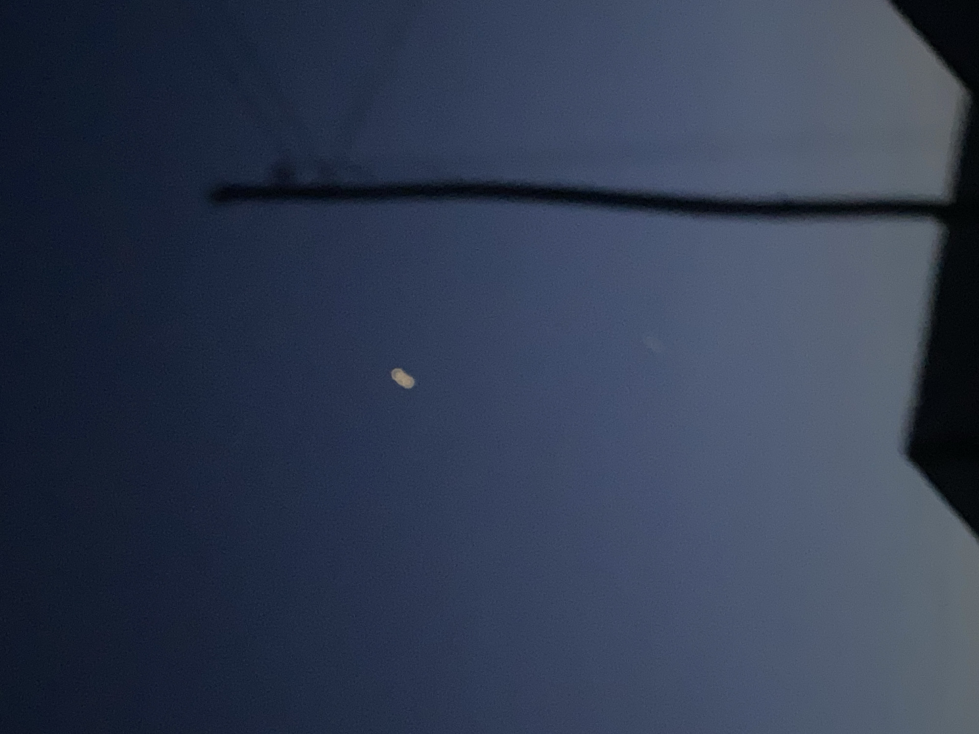 An image taken at dusk in Jalingo, Nigeria, shows a dimly lit scene with a dark blue sky. A slender, vertical pole, likely a utility pole, stands prominently in the right half of the frame, its top extending towards the top edge. A faint, crescent-shaped object, possibly the moon or a distant light, is visible in the upper left quadrant of the sky. The silhouette of a building's roofline is discernible at the bottom of the image, its dark form contrasting with the fading light. The overall impression is one of a quiet, early evening in an urban or suburban setting.