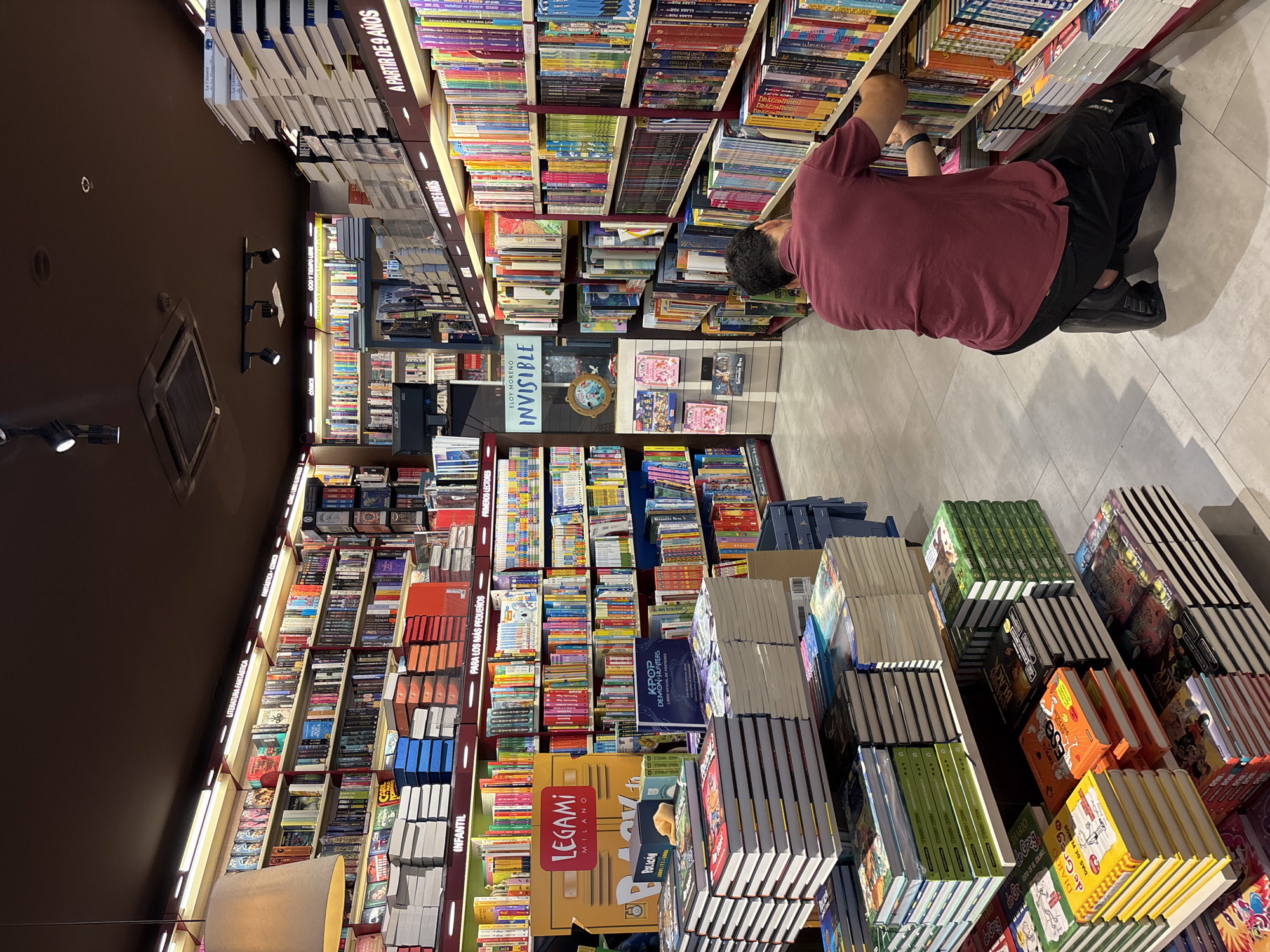 The image depicts the interior of a brightly lit bookstore, likely located in Orihuela, Spain, as indicated by a sign in the background. The store features numerous shelves stocked with books of various sizes and colors, organized into different sections. In the foreground, a man wearing a maroon polo shirt and black pants is kneeling and appears to be organizing or selecting books from a shelf. Stacks of books are visible on the floor and on lower shelves, suggesting a busy retail environment. The lighting comes from overhead fixtures and strip lights along the shelves, illuminating the wide selection of literature available. The overall impression is one of a well-stocked and organized establishment catering to book enthusiasts.