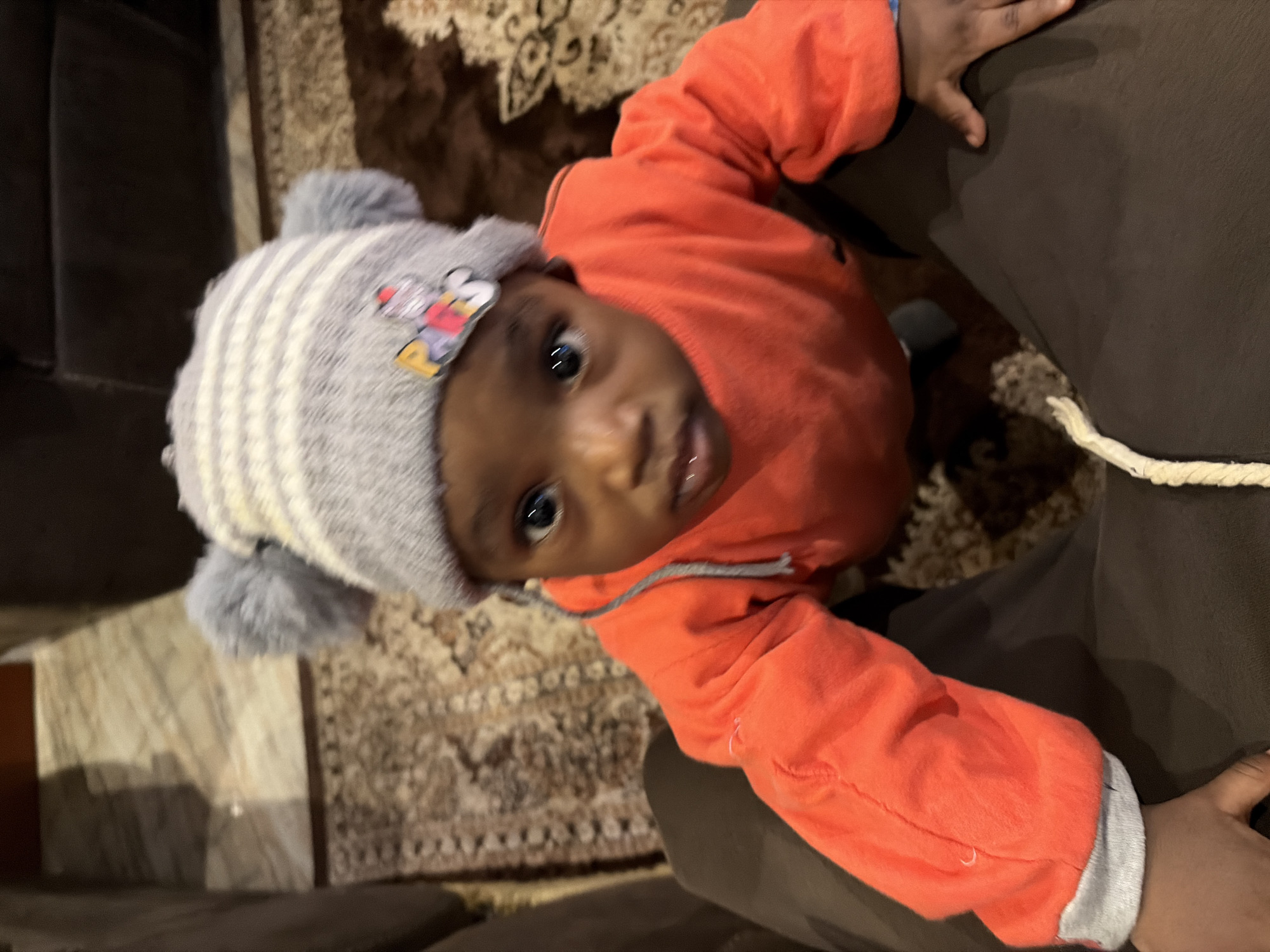 The media file shows a close-up, high-angle shot of a young child wearing a grey and white striped hat with two pom-poms, and an orange hoodie. The child has dark skin and is looking up towards the camera with large, dark eyes and a slightly open mouth. A Mickey Mouse patch is visible on the child's hat. The child is being held by an adult, whose dark-colored pants are visible in the foreground, along with a white rope. The background consists of a patterned rug, suggesting an indoor setting. The lighting appears to be artificial, indicating the photo was likely taken indoors during the day or evening. There is no discernible text in the image. The overall mood is intimate and focuses on the child's expression.