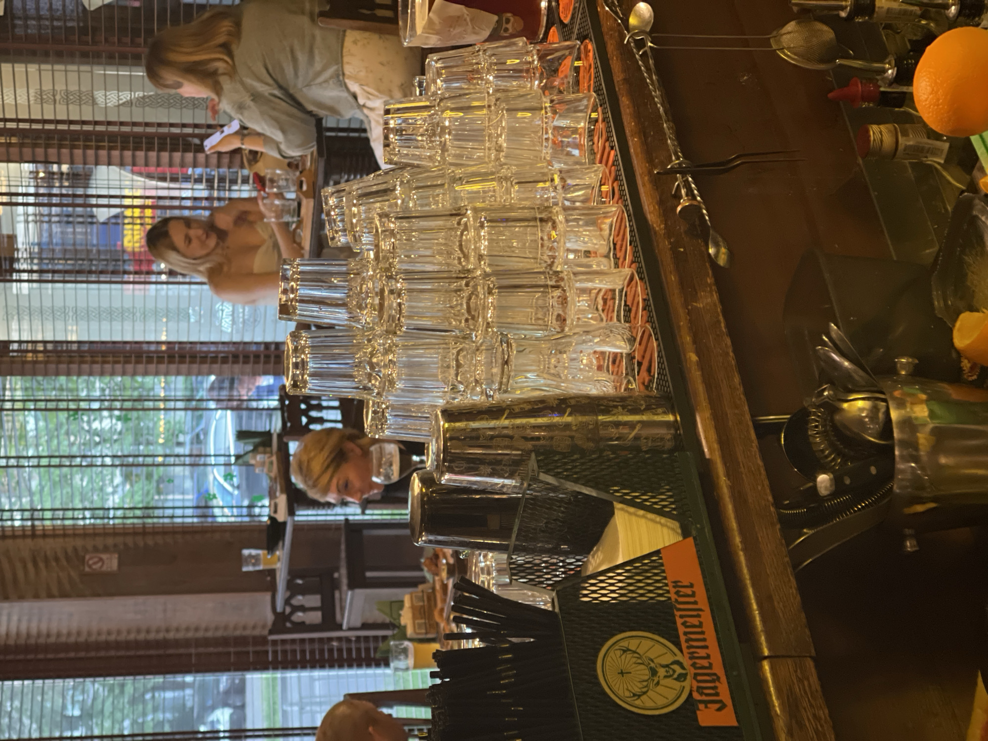 The image depicts the interior of a bar or restaurant, likely in Kyiv, Ukraine, judging by the visible surroundings and potentially the style of establishment. In the foreground, a bar counter is laden with numerous stacks of clean, empty shot glasses. To the left, a black metal holder contains several black straws, and a Jägermeister logo is prominently displayed on the front of the counter. Behind the bar, a variety of bar tools, including shakers and strainers, are visible.
In the mid-ground, several patrons are seated at tables. A woman with blonde hair is facing to the right, looking away from the camera. To her right, another woman with blonde hair is looking down at her smartphone, which she is holding in her left hand. Further back, a woman with blonde hair is visible from the side, wearing a white top. The background shows a window with blinds, through which trees and a street scene are dimly visible, suggesting an outdoor environment beyond the establishment. The overall atmosphere appears relaxed and casual.