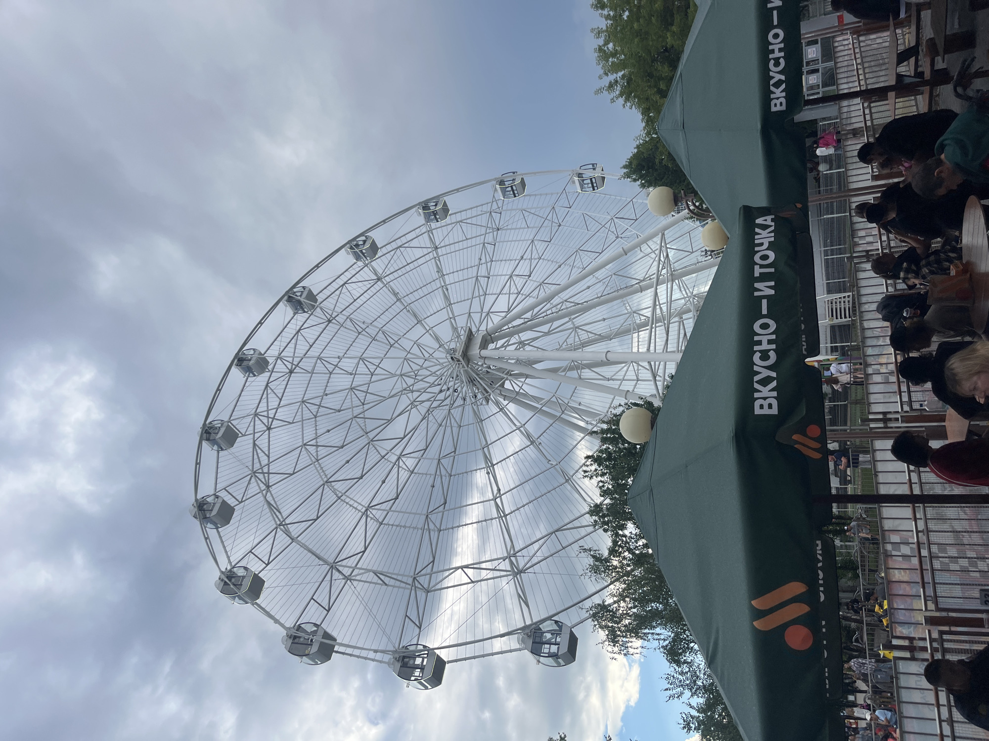 This image shows an outdoor scene at an amusement park. In the background, a large white Ferris wheel with enclosed cabins is prominently displayed against a cloudy sky. In the foreground, two green umbrellas with orange logos and Russian text that reads "ВКУСНО – И ТОЧКА" (Vkusno – i tochka, meaning "Tasty – and that's it") are set up. Several people are seated at tables under the umbrellas, suggesting an eating or resting area. Trees are visible behind the umbrellas and to the left of the Ferris wheel. The overall impression is that of a recreational area on an overcast day. This particular Ferris wheel is located in the city of Tyumen, Russia, at the Tyumen Embankment.