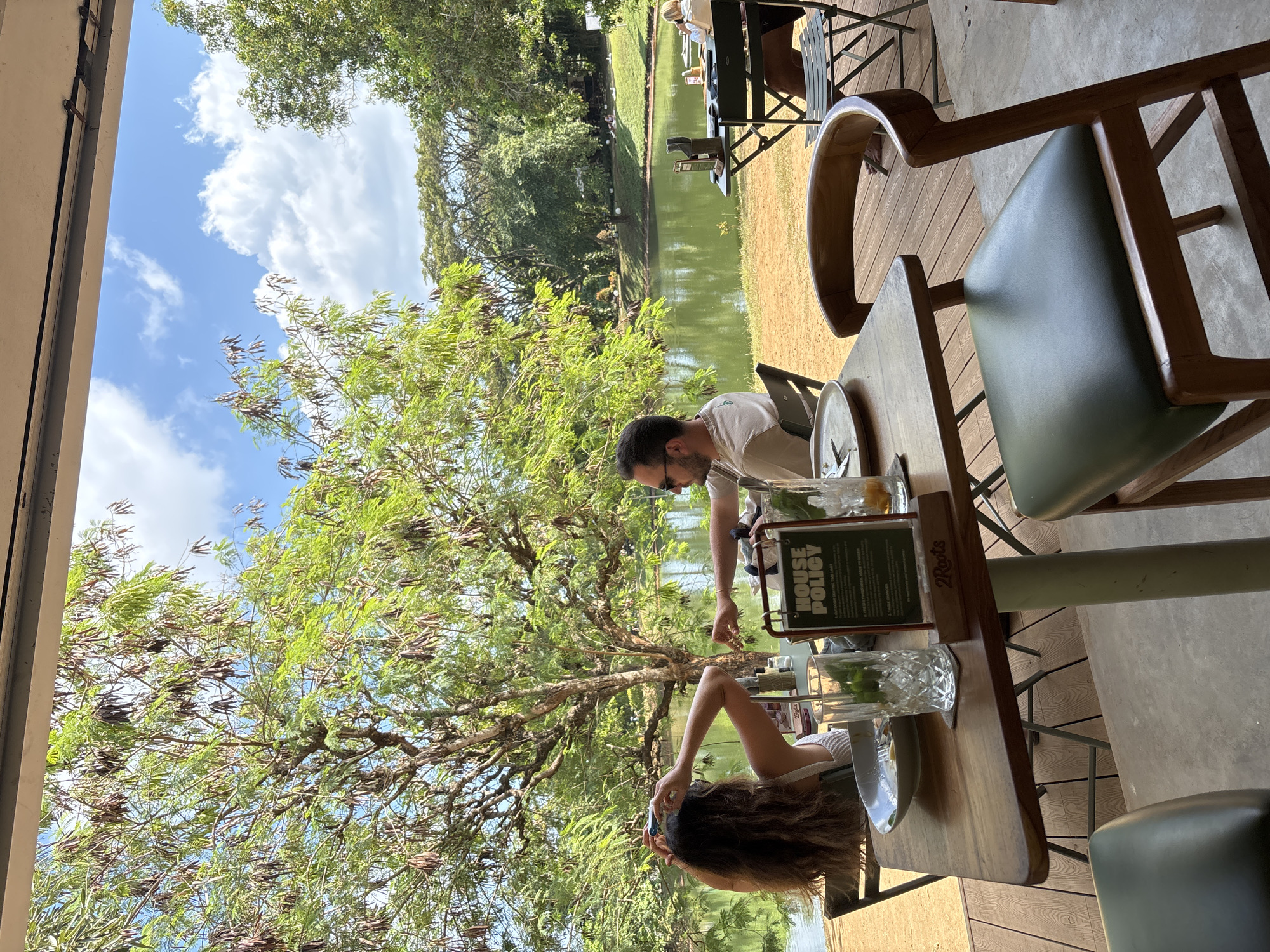 The image captures an outdoor dining experience in Kuta Selatan, Indonesia, on a clear, sunny day with a blue sky dotted with white clouds. The scene is set on a wooden deck, part of what appears to be a restaurant or cafe, overlooking a serene body of water, likely a pond or lake, surrounded by abundant green foliage and large trees.
In the foreground, a wooden table is occupied by two people. A man, wearing sunglasses on his head, is seated and reaching for an item on the table. Opposite him, a woman with long, dark, wavy hair is holding a smartphone, possibly capturing the scenic view or engaging with her device. The table is set with white plates, and two ornate glasses containing drinks with visible mint leaves, suggesting mojitos or similar beverages. A menu stand displays the words "HOUSE POLICY," and a wooden caddy on the table is etched with "2Roots." The chairs feature wooden frames and dark green upholstered seats.
A prominent tree with vibrant green leaves and numerous brown seed pods stands between the dining area and the water, its branches extending across the frame. Beyond the water, a distant shore is visible with more dense tree lines and hints of open space or grass, suggesting a tranquil and natural environment. The overall impression is one of leisure and enjoying a meal or drinks in a picturesque, open-air setting.