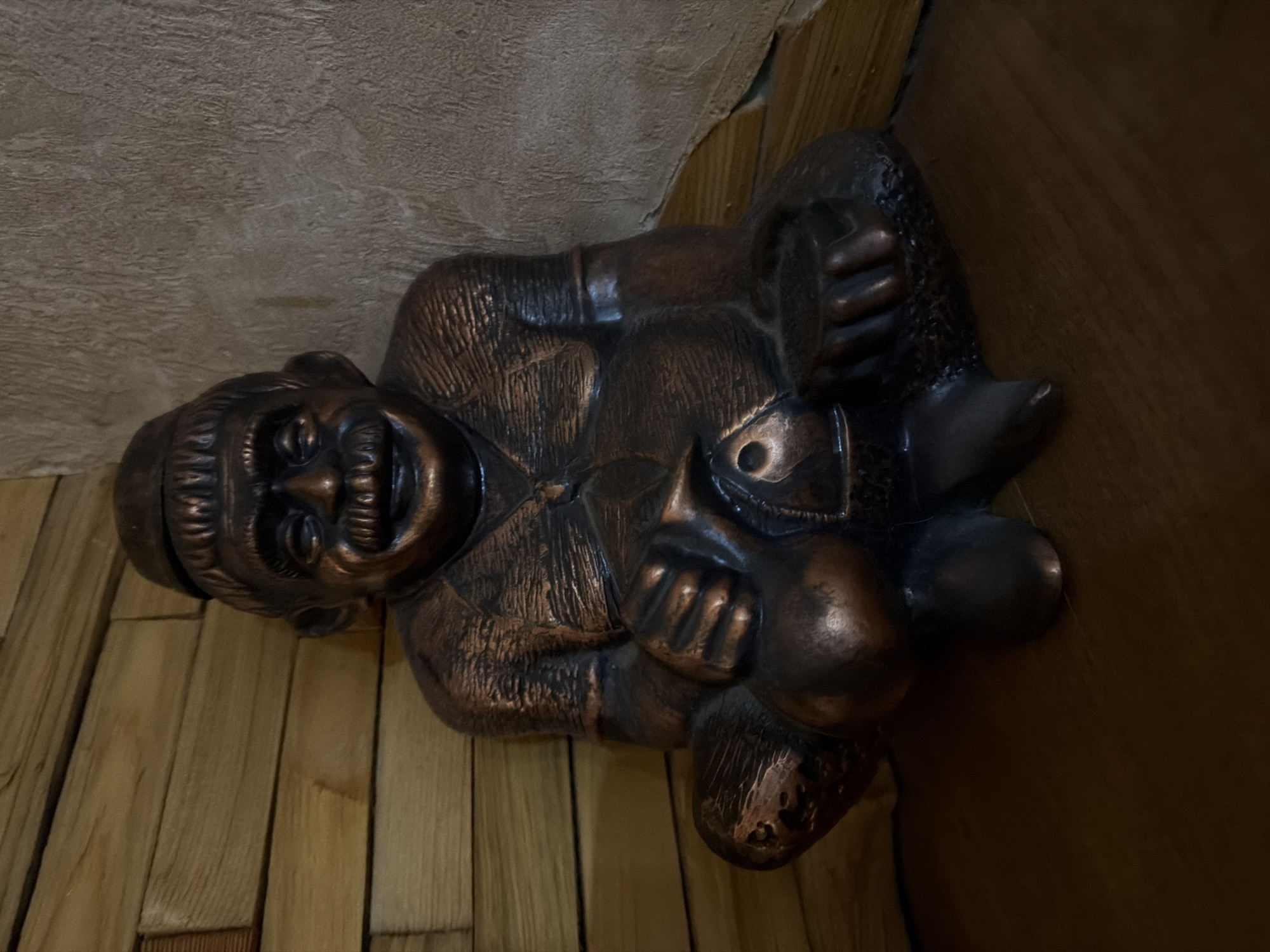 A dark bronze statue of a man with a large mustache and a cap sits on a wooden floor against a wall. The wall is partially paneled with horizontal wooden planks and partially textured with a rough, tan material. The statue is in a seated position with its legs crossed and its hands resting on its lap. It appears to be holding a small fish in its left hand. The lighting is dim, creating shadows on the statue and the surrounding environment. The statue's expression is one of amusement, with wide eyes and a broad smile.