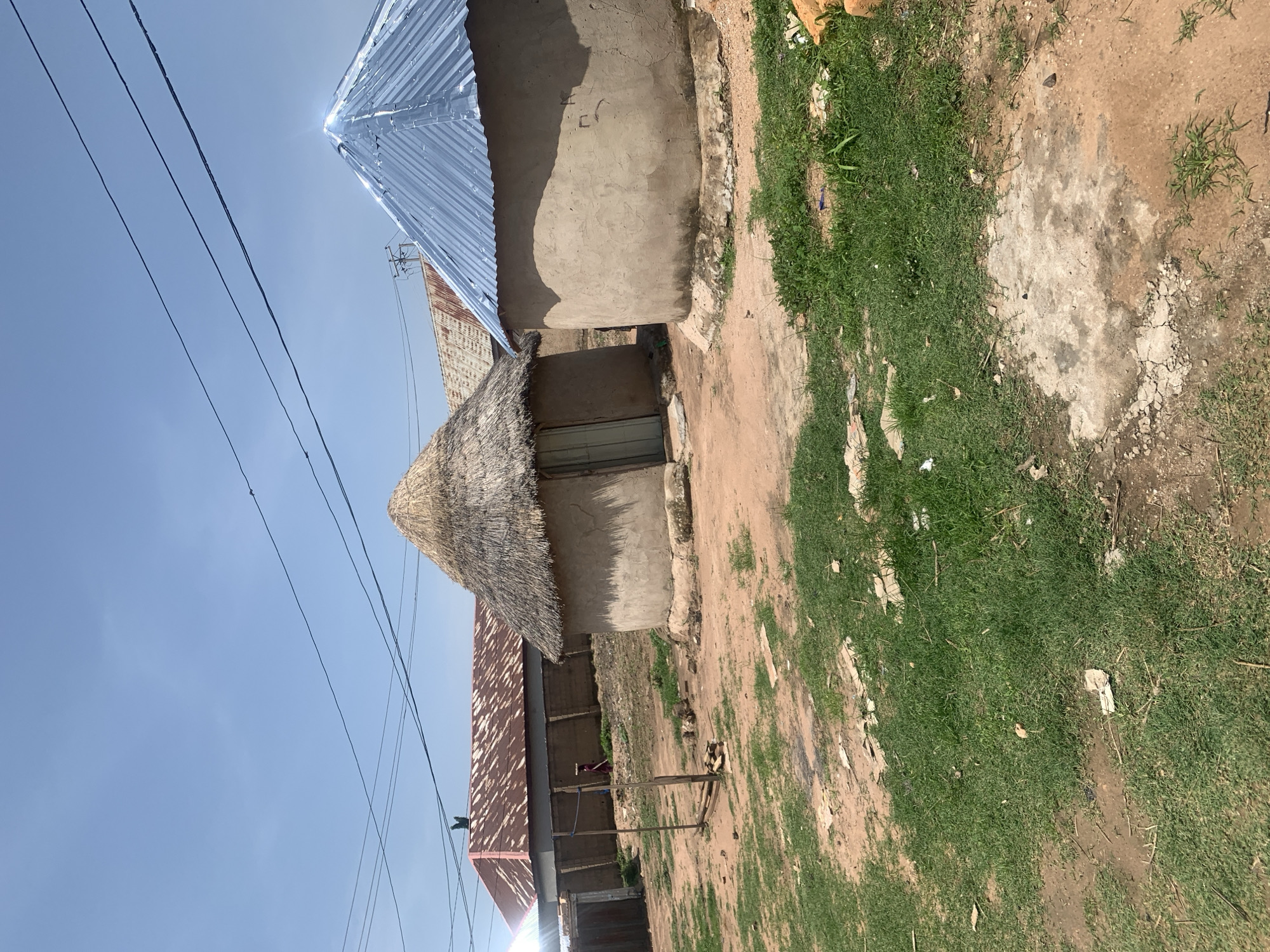 This image depicts a scene in Jauro Yinu, Nigeria, featuring traditional round dwellings with thatched and corrugated metal roofs. In the foreground, the ground is a mixture of dirt and patches of green grass. To the left, a fence encloses a compound with a wooden door. Two round huts are prominent: one with a thatched roof and a light-colored door, and another to its right with a shiny blue corrugated metal roof. In the background, a larger building with a rusty corrugated metal roof is visible, and a person in a dark red garment is walking near it. Power lines crisscross the bright blue sky above. The overall environment suggests a rural or semi-urban setting in Nigeria.