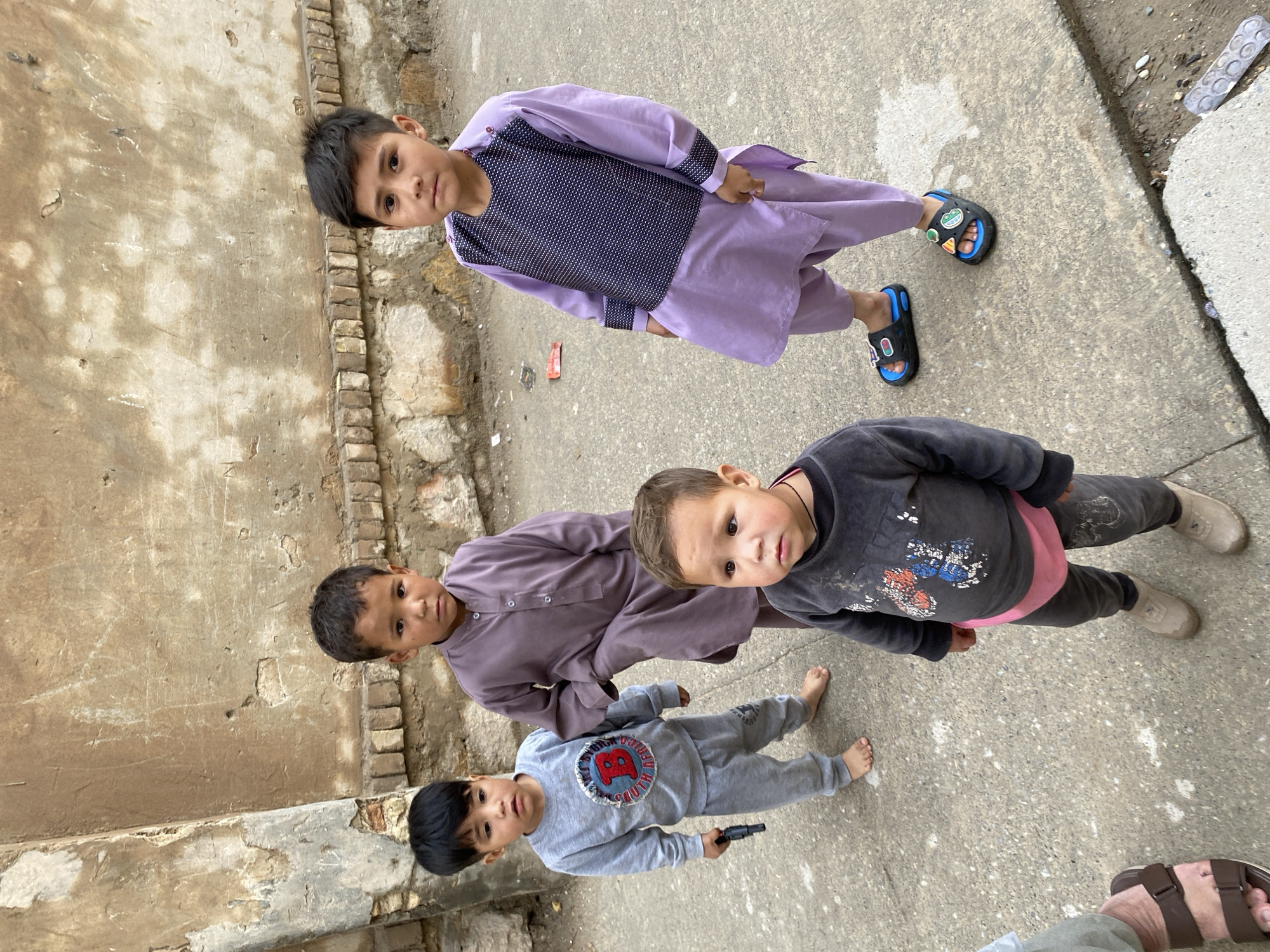 The image displays four young male children standing on a grey concrete surface against a weathered, light brown wall in Kandahar, Afghanistan. The lower portion of an adult's feet, wearing brown sandals, is visible in the bottom-left foreground.
From left to right, the children are arranged as follows:
1. A male, estimated 3-5 years old, wears a grey long-sleeved top with a "B SOUTH AFRICA" emblem and matching grey sweatpants. He is barefoot and holds a dark-colored toy pistol in his right hand. He looks slightly to the left of the camera.
2. A male, estimated 4-6 years old, wears a plain long-sleeved purple kurta-style top and matching trousers. He is barefoot and looks directly at the camera.
3. A male, estimated 2-4 years old, stands in the immediate foreground, wearing a dark grey long-sleeved top with multi-colored paint or dirt marks and dark grey trousers. A pink undergarment is visible at his waist. He wears light brown, low-cut shoes and looks slightly to the left of the camera.
4. A male, estimated 4-6 years old, wears a light purple long-sleeved kurta with a dark blue patterned chest panel and matching cuffs, along with light purple trousers. He wears black and blue slide sandals adorned with several small decorative patches. He looks directly at the camera.
The background wall exhibits significant signs of age, including chipped plaster, exposed bricks, and cracks. The concrete ground shows scattered light stains or marks, and minor refuse such as a red wrapper and a white plastic blister pack are visible near the wall on the right.