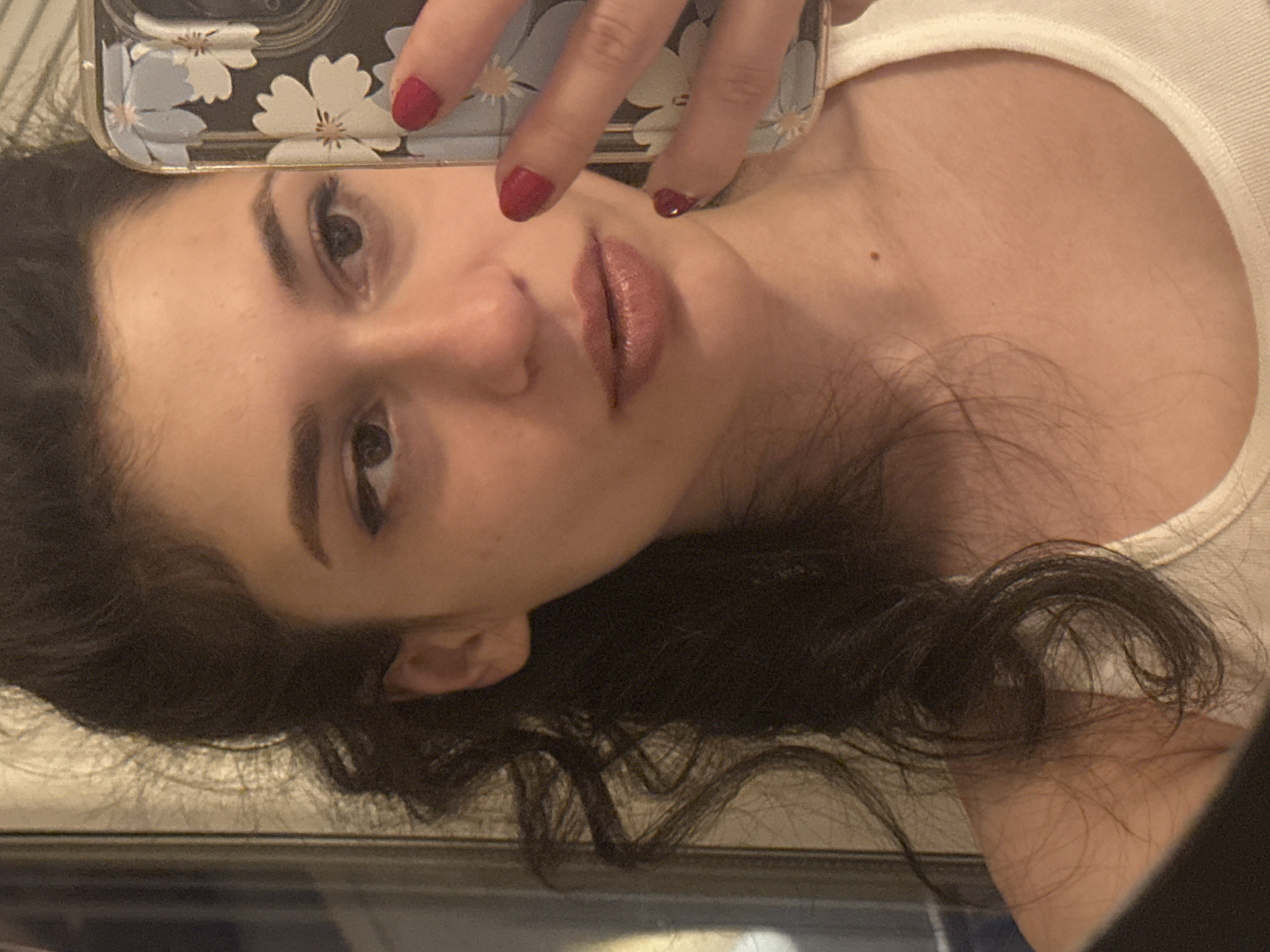 A close-up selfie is captured from the chest up, focusing on a young woman's face and upper body. She has dark, curly hair pulled back into a ponytail, with some strands framing her face. Her skin appears smooth, and she is wearing subtle makeup, including eyeliner and lipstick. Her left hand, adorned with red nail polish, is holding a smartphone with a floral case, positioned to take the picture. The background is blurred, but hints of a bathroom setting are visible, with what appears to be a mirror and tiled wall. The lighting is warm and soft. The image does not provide any information about the specific location within Sofia, Bulgaria, or any details about the surroundings beyond the immediate, personal space.