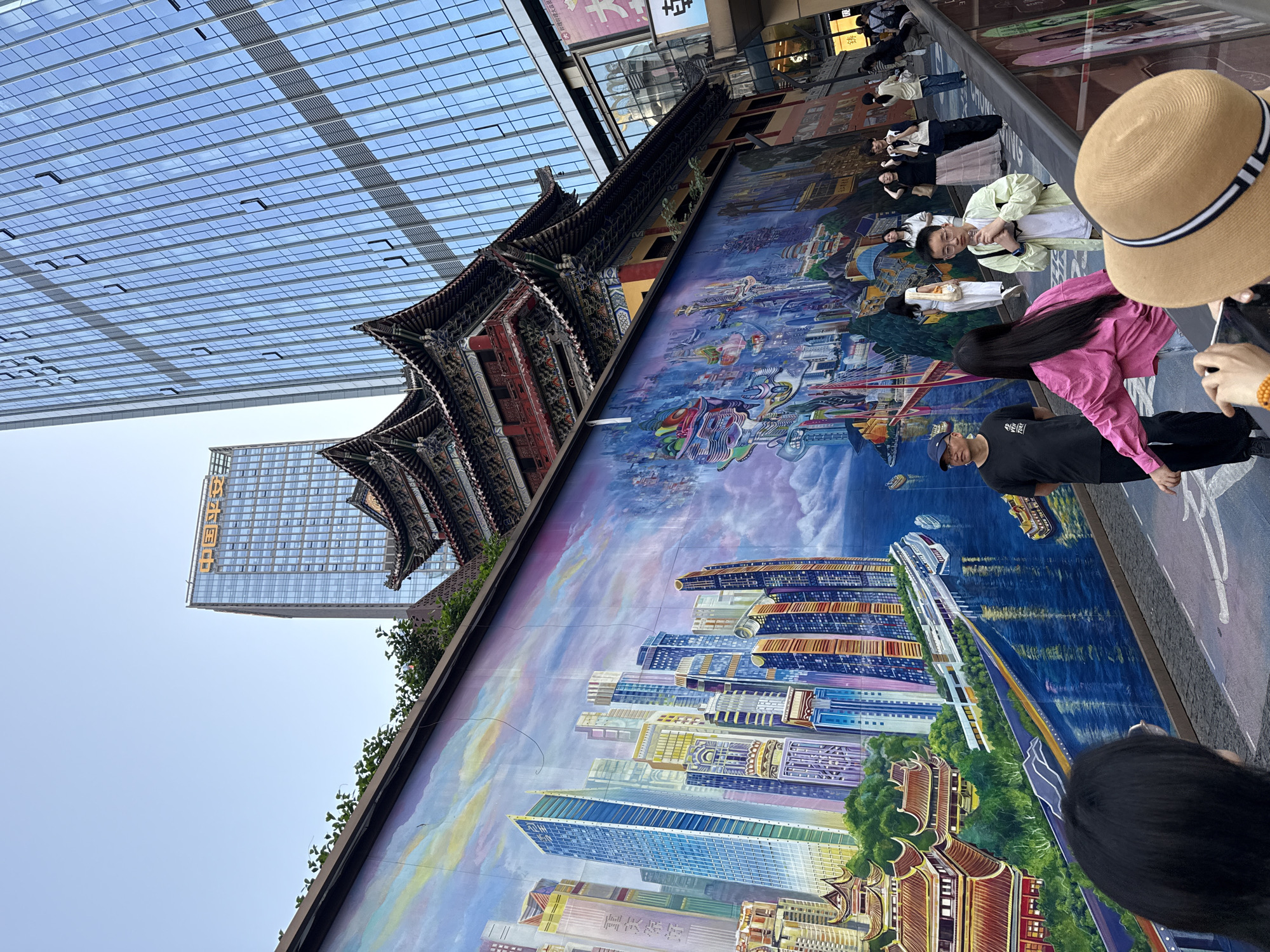 The image shows a mural in Yuzhong District, China, that blends a traditional Chinese temple style with modern skyscrapers. The artwork is set on a city plaza floor where people are standing and interacting with the art. In the background, one can see a modern glass-walled high-rise building, juxtaposing the old and new themes in the artwork. People are viewing and posing with the mural, with one person wearing a straw hat and another wearing a pink jacket. Other individuals are casually dressed and engaged in taking pictures or observing. The scene is outdoors with clear weather.
