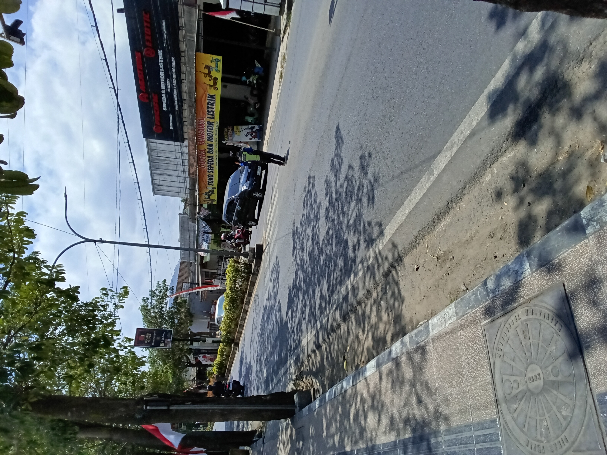 The image depicts a street scene in the city of Kelutan, Indonesia. The scene is characterized by a road with white lane markings, bordered by a paved sidewalk on the left. Trees with dappled shade from sunlight cast shadows across the road and sidewalk. On the right side of the road, there are buildings with storefronts and banners. One banner prominently displays "PUTRA WIBAWA TOKO SEPEDA DAN MOTOR LISTRIK," indicating a shop selling electric bicycles and motorcycles. Another large black sign above this banner advertises "PACIFIC EXOTIC SEPEDA & MOTOR LISTRIK," further emphasizing the presence of electric vehicle sales and service.
In the middle of the road, a person wearing a reflective vest and a dark uniform is standing, appearing to be directing traffic. A dark-colored van is stopped in front of this individual, and various motorcycles are visible on the side of the road. A red and white Indonesian flag is mounted on a pole on the left side of the street, and another is visible on the right, suggesting a sense of national presence. The sky is partly cloudy, with a bright, clear blue sky visible between the clouds. The overall impression is a bustling commercial street with ongoing activity.