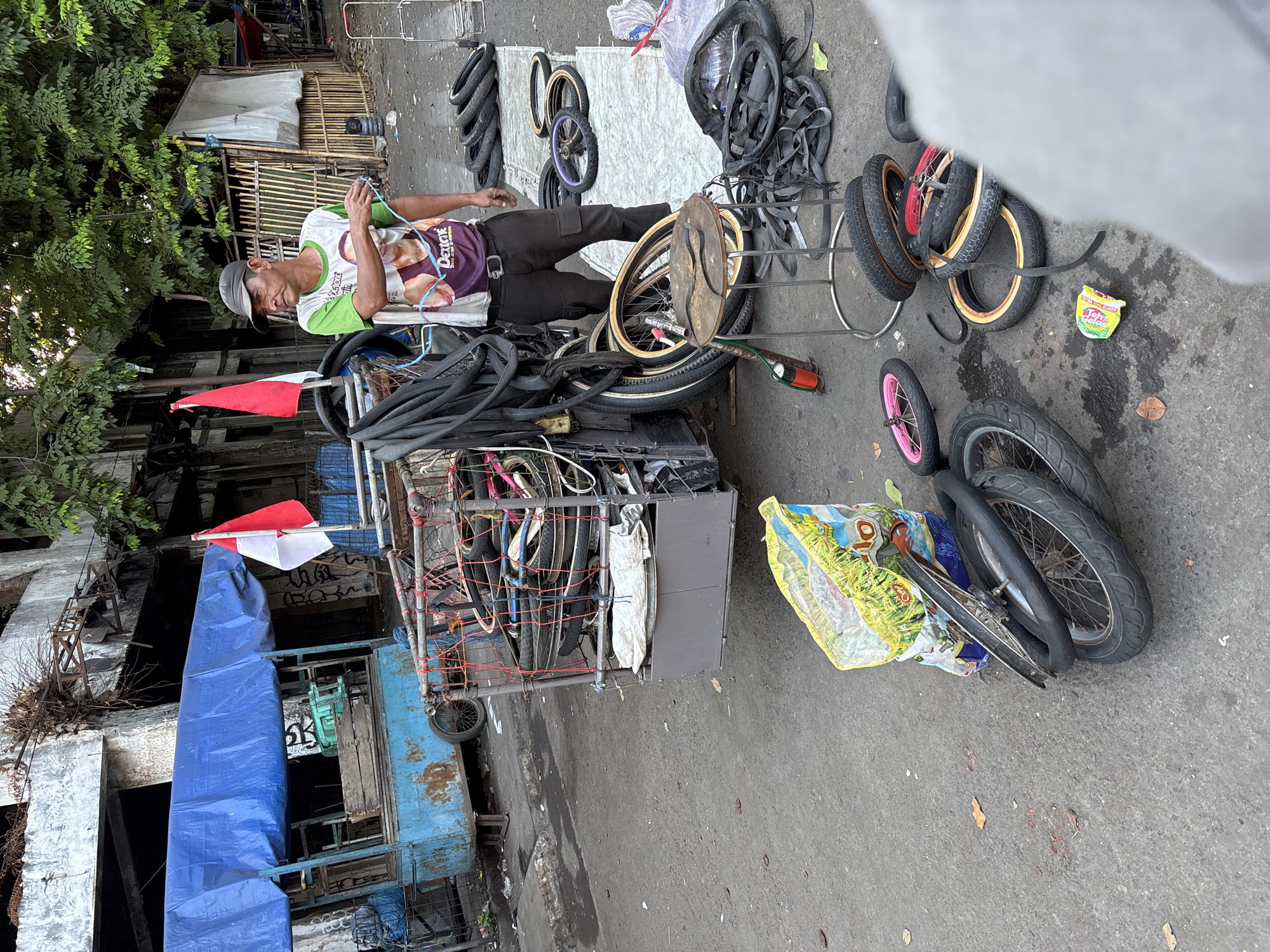 The image depicts an outdoor street scene in Cirebon, Indonesia. A man wearing a cap and a t-shirt with images of two men on it stands near a mobile cart laden with bicycle and motorcycle tires and inner tubes. He appears to be a vendor, possibly a tire repairman. The cart is made of metal and has a blue tarp covering part of it. In the background, there are Indonesian flags displayed, indicating a patriotic presence. The environment shows signs of urban decay, with an abandoned or dilapidated building visible behind the cart. The ground is paved with asphalt, and there are various discarded tires and other items scattered around. The scene suggests a bustling, informal economy operating in a less-than-pristine urban setting.