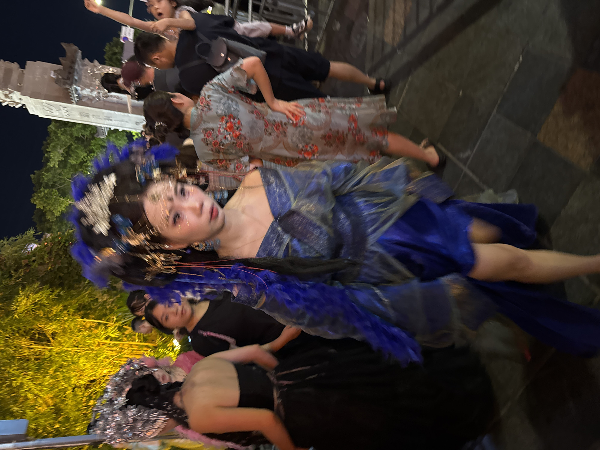 In the Yuzhong District of China, at night, a woman in a blue and gold elaborate costume and headdress walks towards the viewer. She has dramatic makeup on her eyes and forehead and her hair is dark. The costume features blue feathers and flowing fabric. Behind her, a crowd of people are visible, some dressed in casual attire and others in costumes. To the right of the frame, a large stone monument is partially visible. The lighting suggests an outdoor event, possibly a festival or parade, with illuminated trees in the background.