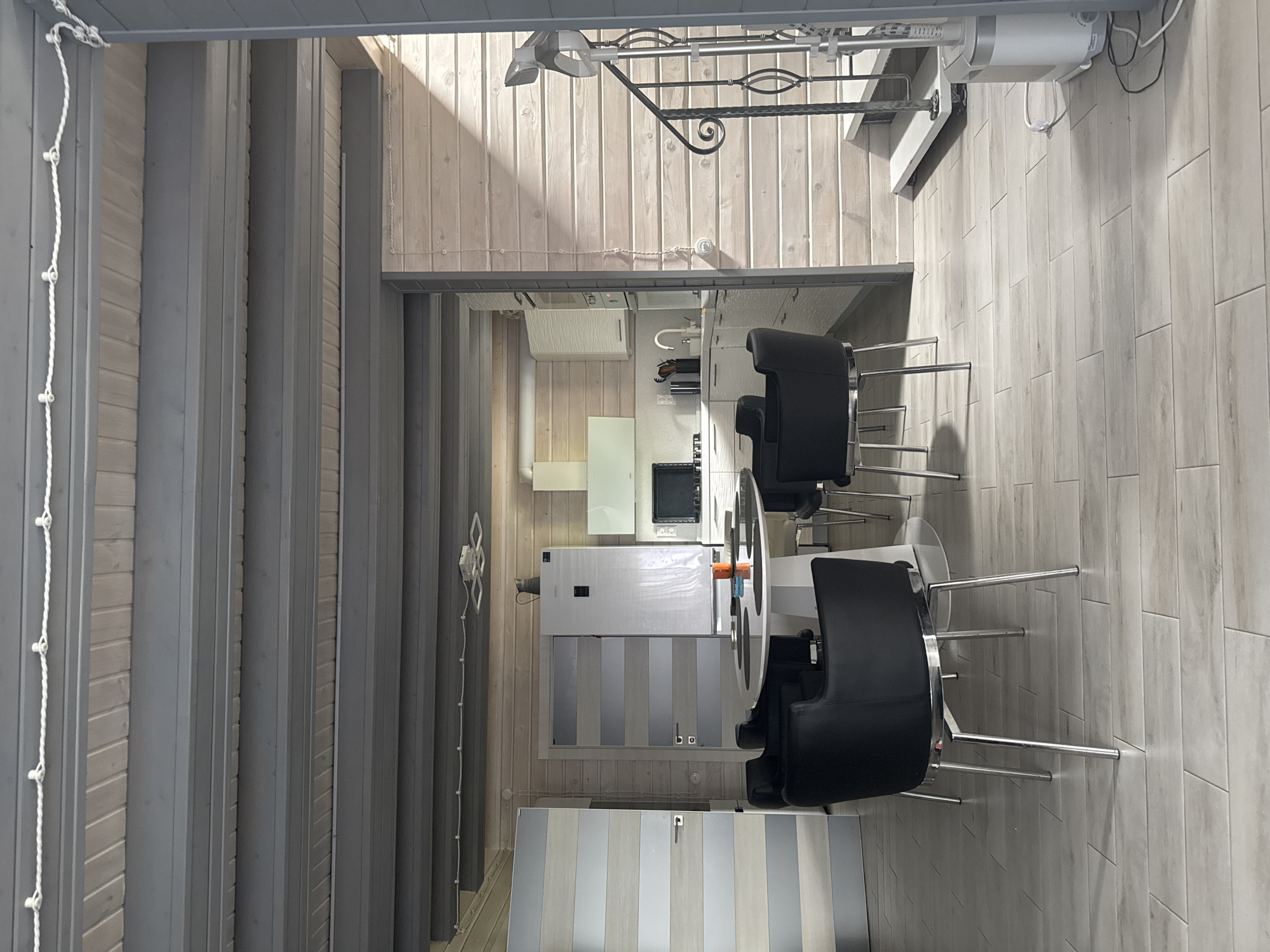This image shows an interior view of a room in a house, possibly a dining and kitchen area, located in Monino, Russia. The room features a white round dining table with four black chairs around it. To the left, there is a door with horizontal stripes of wood and silver. Beyond the dining area, a kitchen is visible with white cabinets, a refrigerator, a stovetop, and a sink. To the right, a staircase with a black metal railing leads upwards, and a white upright appliance is situated at the bottom of the stairs. The walls are paneled with light-colored wood, and the ceiling has exposed gray beams. Natural light streams in from the right side of the image, illuminating the wooden walls and creating shadows. There are no people visible in the scene.