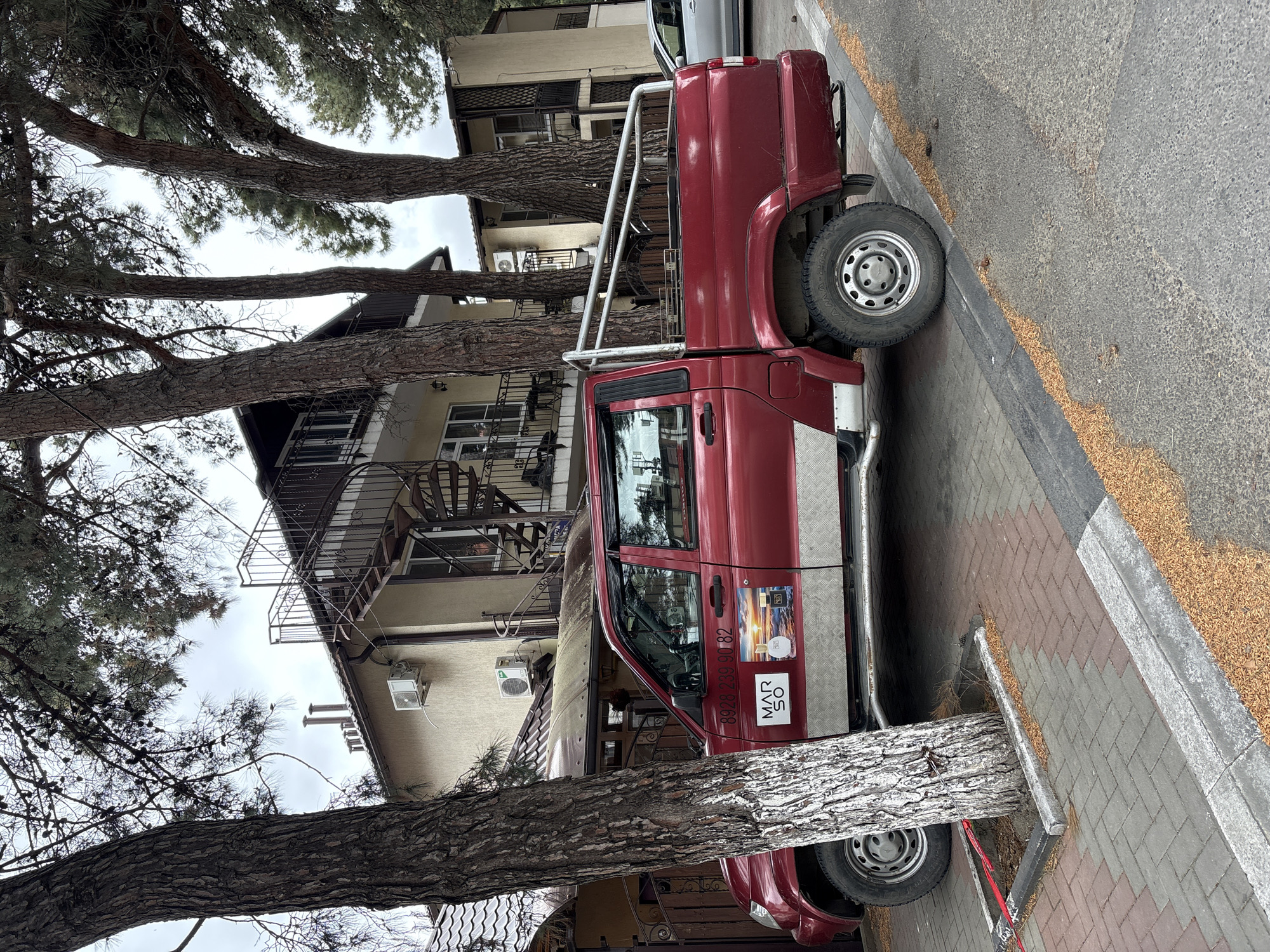 A red pickup truck is parked on the side of a brick-paved street in Gelendzhik, Russia, as indicated by the "MAR 50" sticker with a Russian phone number on the driver's side door. The truck has a chrome running board and ladder rack. A multi-story building with a beige exterior is visible in the background, featuring an external spiral staircase. The scene is partially obscured by mature trees, their trunks positioned between the vehicle and the camera, adding foreground elements to the image. The sky is overcast.
