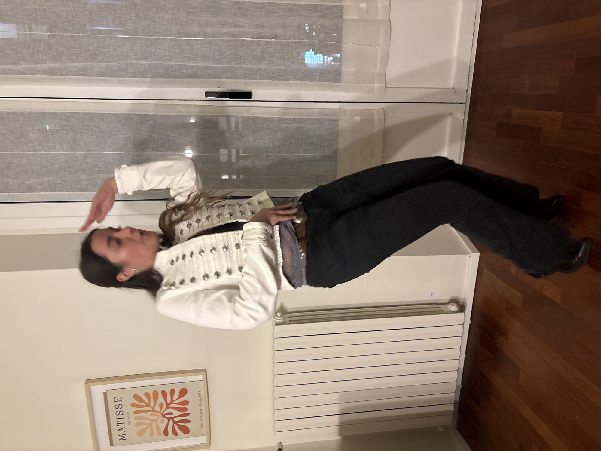 A person is standing on a window seat in front of a window. They are wearing a white jacket with silver embellishments and dark pants. They are holding their hand up to their forehead, as if shading their eyes. There is a radiator below the window seat and a framed Matisse print on the wall to the left. The scene is indoors, likely a residence, with hardwood floors visible. Through the window, an urban landscape is visible, suggesting that the location is in Milan, Italy.