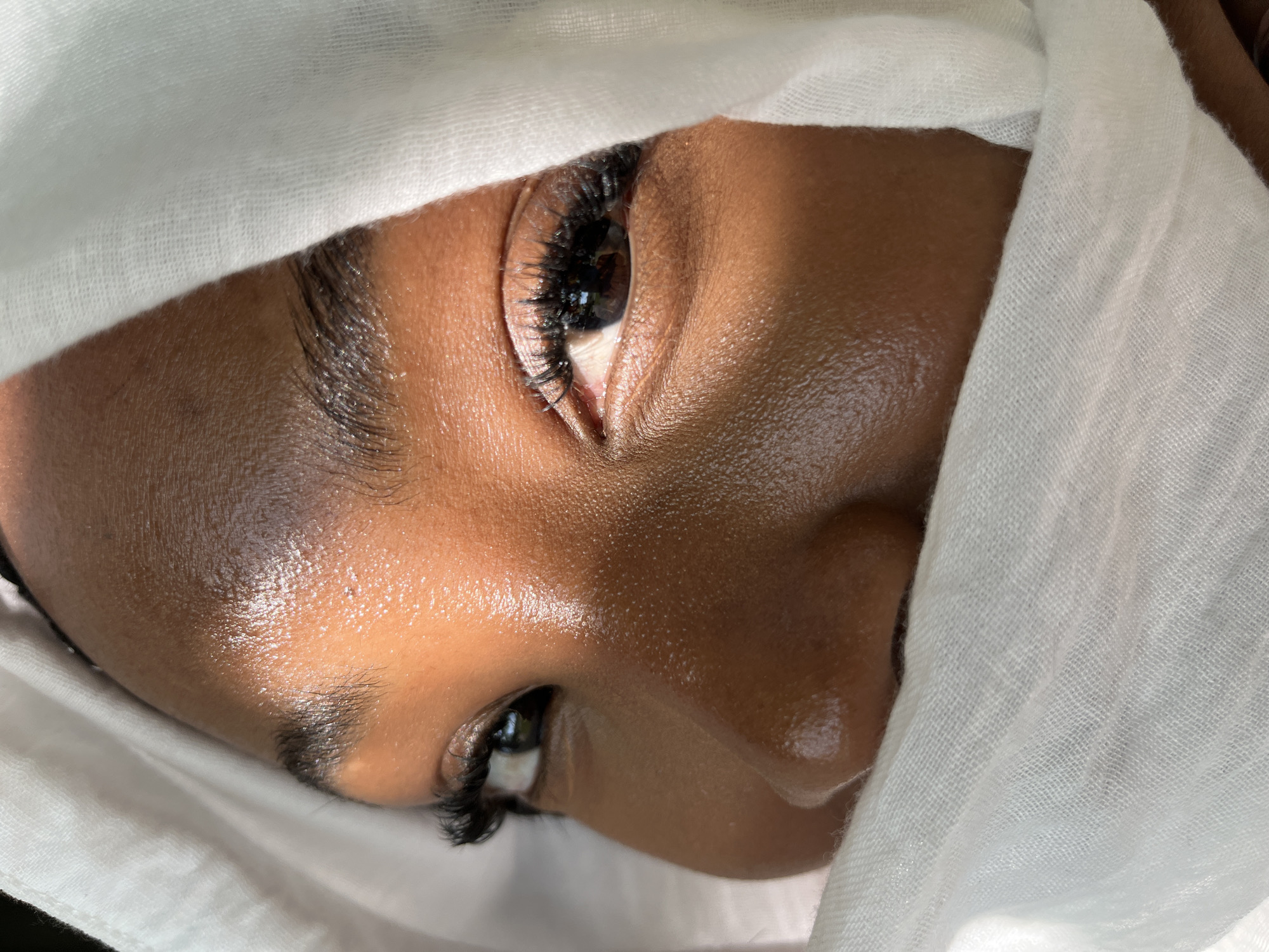 A close-up, eye-level shot focuses on the right side of a person's face, from the nose up to the forehead, wrapped in a sheer white fabric. The fabric drapes over the forehead and partially obscures the nose and mouth. Sunlight casts a diagonal stripe of light across the forehead, highlighting the texture of the skin, and also illuminates the right eye and cheekbone. The individual has dark eyes with long, dark eyelashes, and visible eyebrows. The skin appears dewy and reflective. The background is indistinct, primarily composed of the white fabric and shadowed areas. There is no information to place this image in Kazan, Russia.