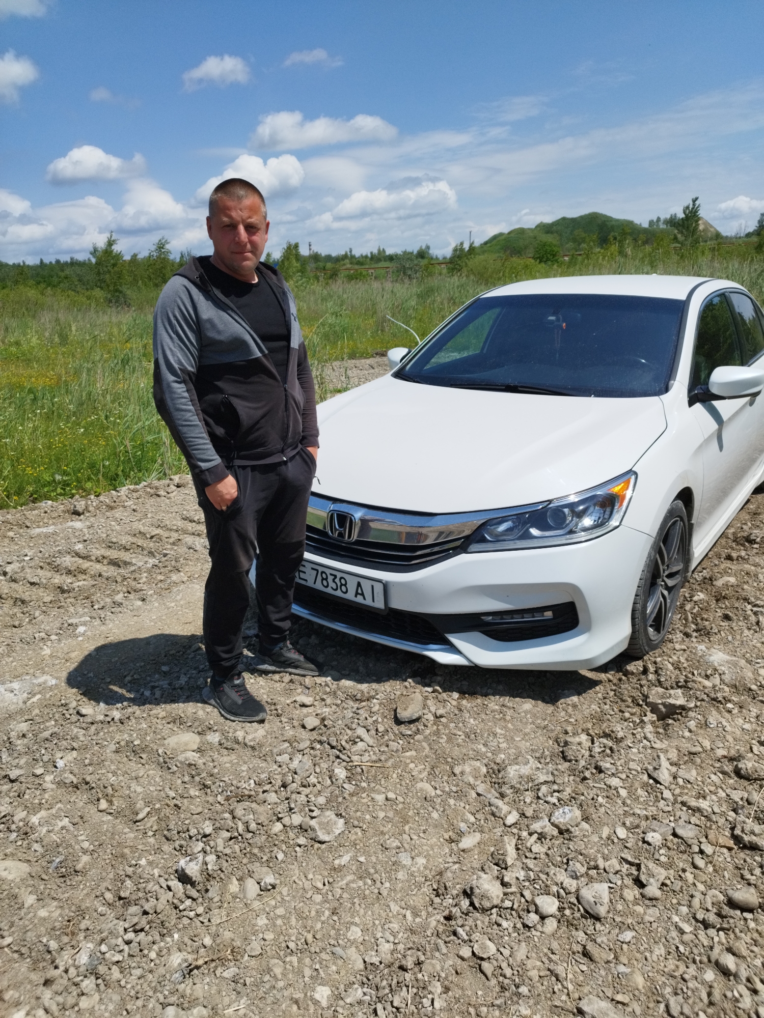 A man stands next to a white sedan in a rural outdoor setting. The man is wearing a grey and black zippered hoodie over a black t-shirt, and black sweatpants with black and grey athletic shoes. He has his right hand in his sweatpants pocket, and his left arm is bent with his hand resting on his hip. He has short hair and is looking at the camera with a neutral expression.
The car is a white Honda sedan. Visible text on the car's license plate reads "E 7838 AI". The car is parked on a dirt and gravel road. Behind the car and the man, a grassy field with scattered green trees and some taller green foliage stretches towards a hazy, tree-covered hill in the background. The sky is blue with scattered white clouds. The scene suggests it is daytime, likely late morning or early afternoon, given the bright sunlight and clear sky. The location cues, such as the rural landscape and the specific license plate format, point to Rozdil, Ukraine. The overall mood is casual and relaxed.