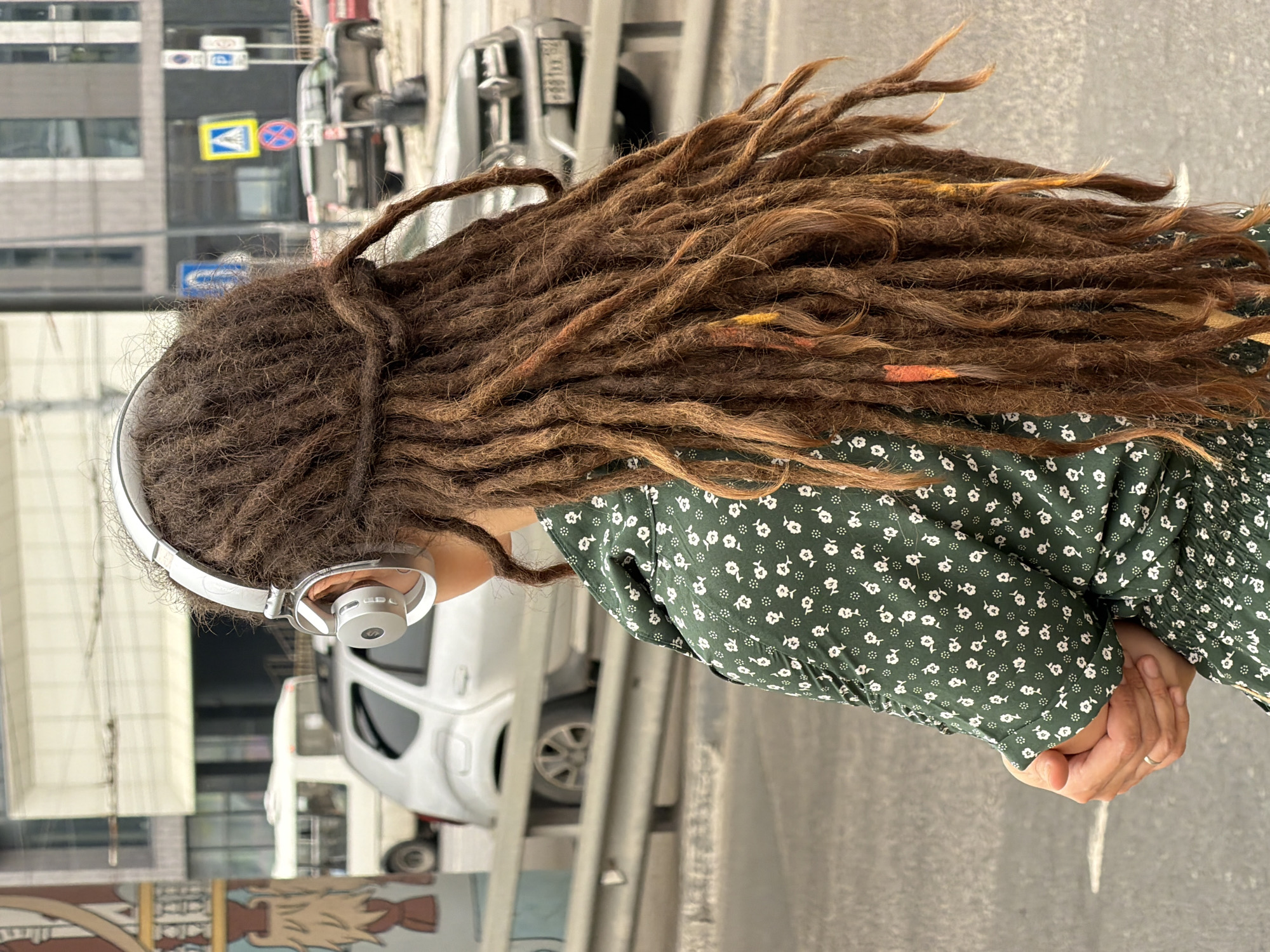 A woman with long, brown dreadlocks, some with orange and yellow streaks, wears silver headphones and a dark green dress with a white floral pattern. She is standing on a street in Nizhny Novgorod, Russia. In the background, there are cars, buildings, and traffic signs. The scene appears to be on an overcast day, with a focus on the woman's back and her distinctive hair.