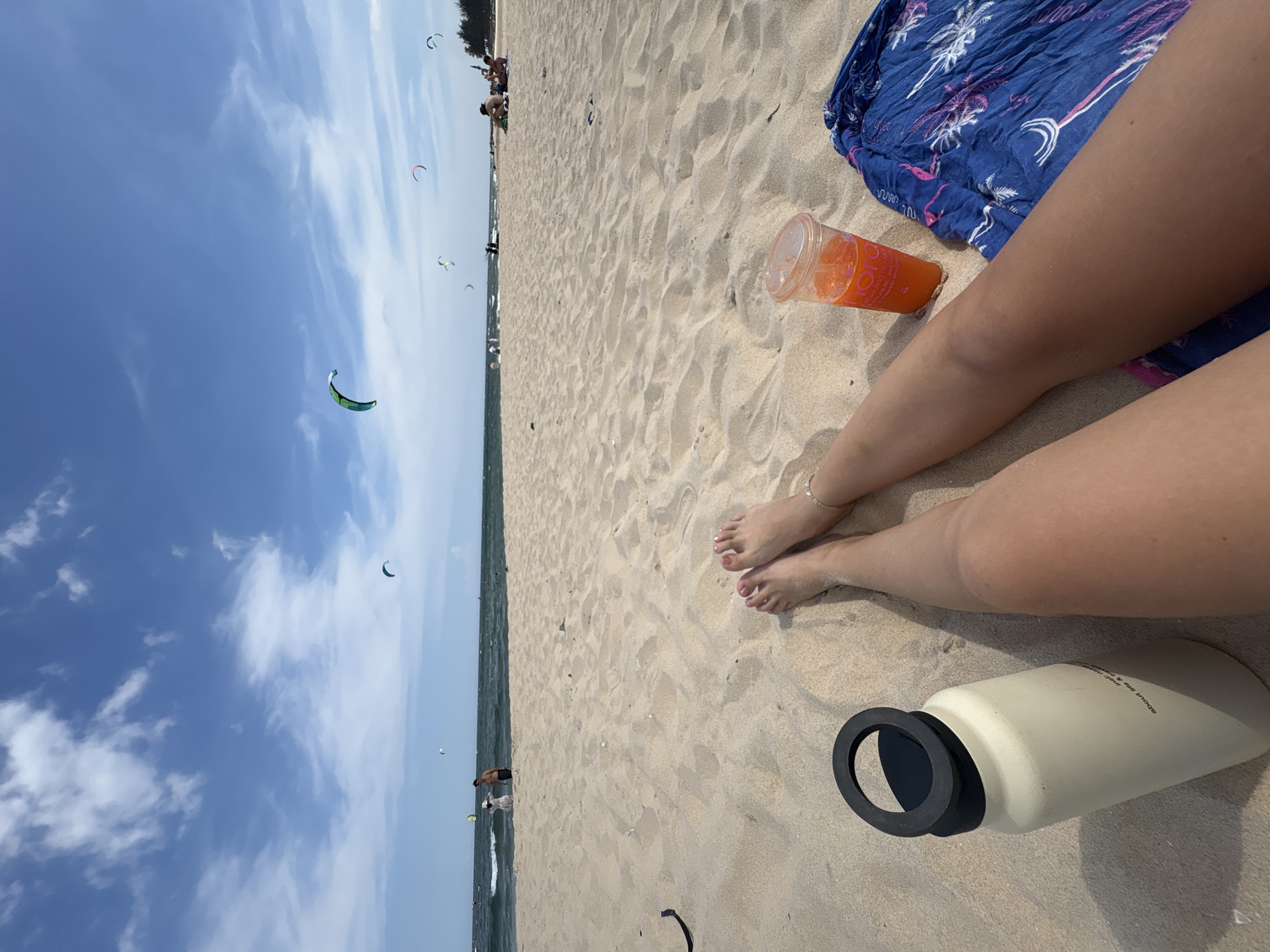 This is a first-person perspective shot from a beach in Mũi Né, Vietnam. In the foreground, the legs of a person are visible, with a light-colored water bottle to the left and a plastic cup of orange juice and a blue patterned sarong to the right. The sand is light and granular. In the middle ground, the ocean is visible with gentle waves and several kitesurfers in the water. The sky is blue with scattered white clouds. In the background, more people are seen on the beach and in the water, and a line of trees is visible in the distance.