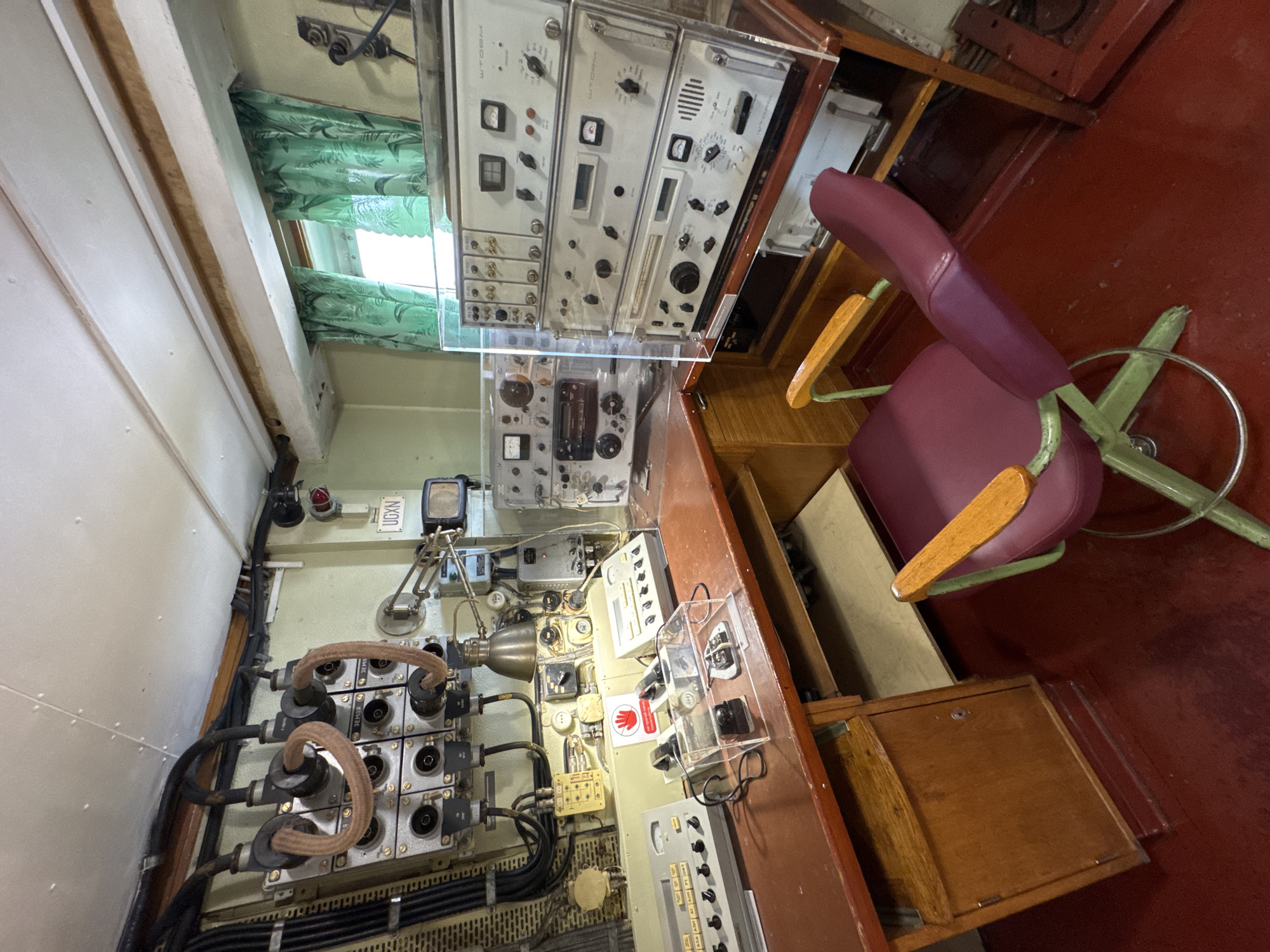 The image shows a room with vintage electronic equipment, possibly a radio room or control center. A wooden desk is placed against a wall, covered with various devices, including oscilloscopes, power supplies, and switches. A red chair with a metal base and armrests is positioned in front of the desk. The room has a green painted wall, a white ceiling, and a red floor. A window with green curtains is visible in the background. There is a sign that reads "UGXN" above a small screen. The overall impression is of a functional space with a retro aesthetic. The image does not provide enough information to determine the specific location in St. Petersburg, Russia.