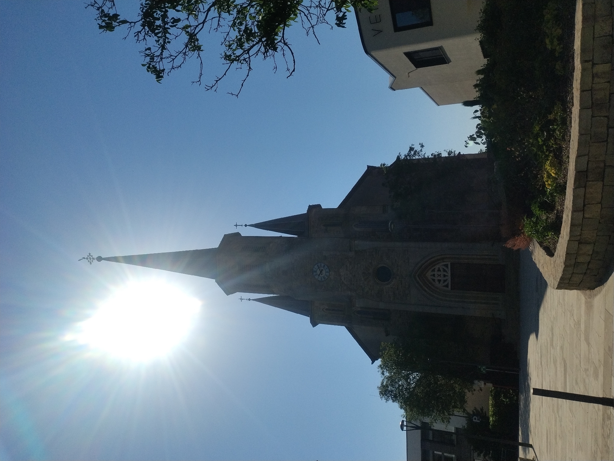 In the city of Crauthem, Luxembourg, a tall stone church with a steeple and a cross on top is captured from a low angle. The sun shines brightly in the clear blue sky, causing a lens flare. The church has a clock face and a large arched doorway with ornate detailing. In front of the church, a paved area leads to a garden with a low stone wall. To the right of the church, a modern building with a white facade and dark windows is visible. A tree with green leaves branches out into the sky on the right side of the image. The scene is set on a sunny day.