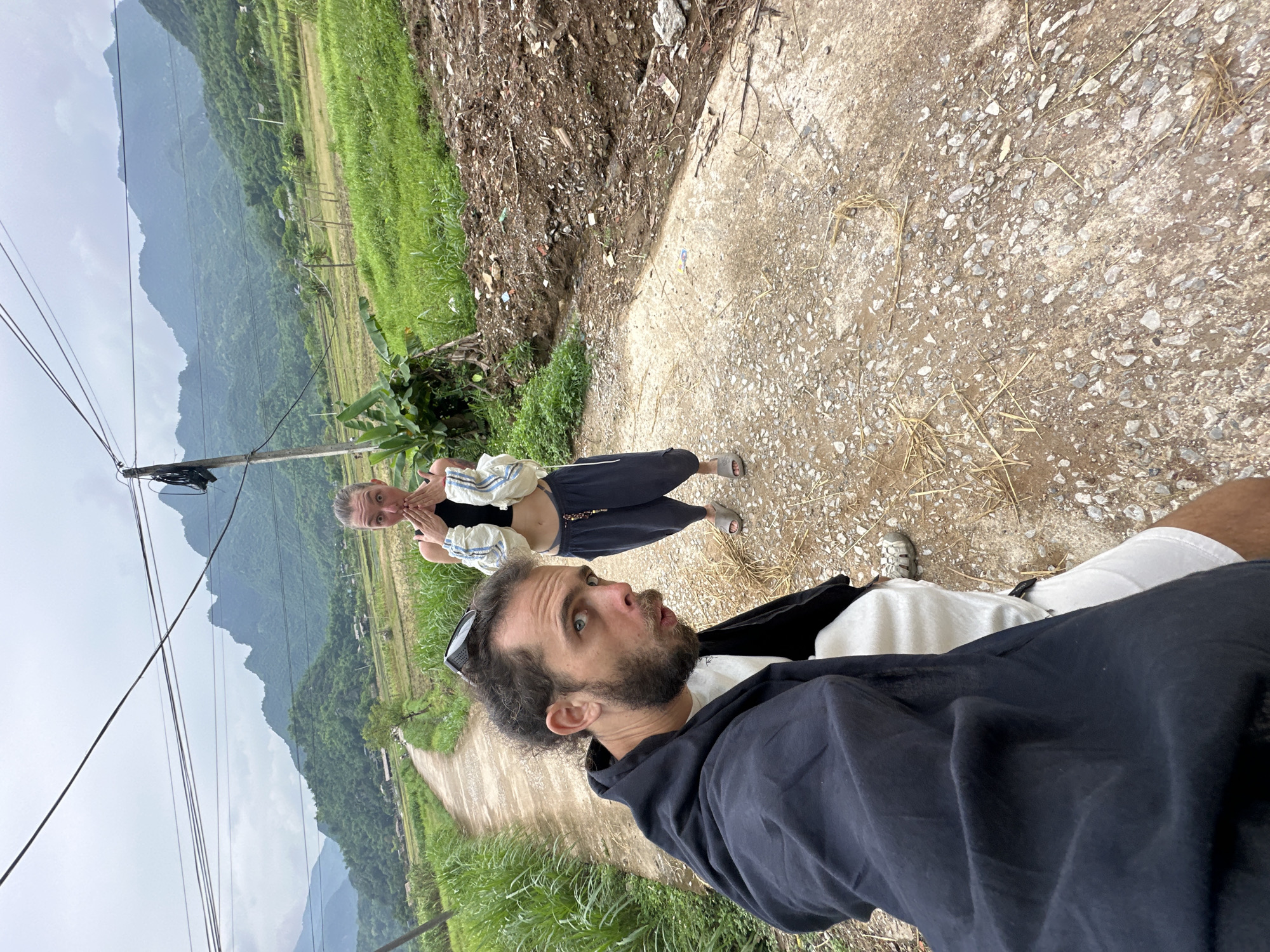A selfie is taken by a man with curly brown hair and a beard, who is looking at the camera with a surprised expression. He is wearing a black hooded jacket over a white t-shirt and white shorts. Behind him, a woman with her hair pulled back is looking at the camera with her hands covering her mouth. She is wearing a black crop top and dark pants. They are standing on a gravel road in a rural area. The road is lined with green grass and vegetation, and in the background, there are terraced rice fields and lush green mountains under a cloudy sky. The location is identified as Mai Chau, Vietnam.