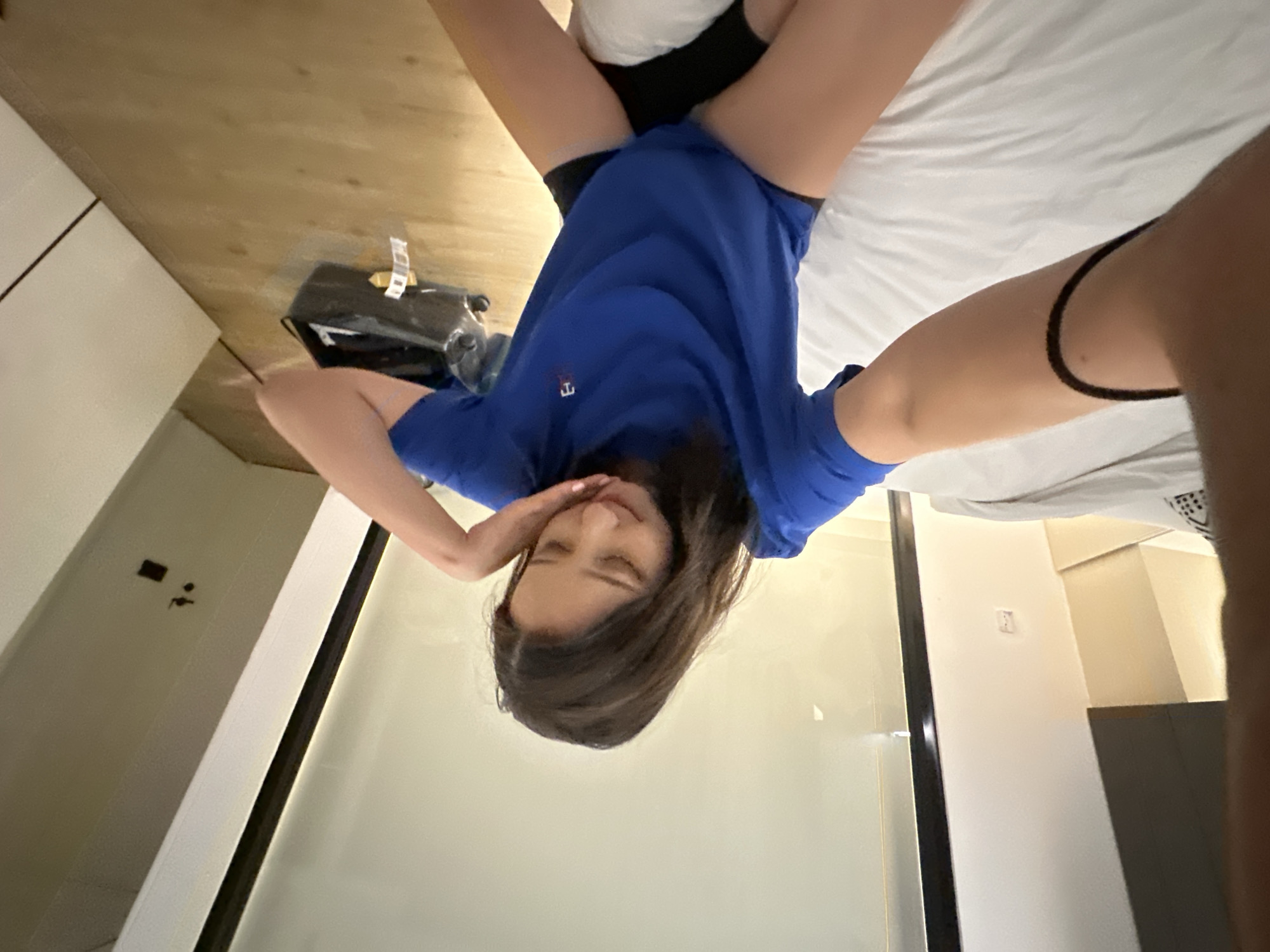 The image shows a young woman with brown hair sitting on a bed. She is wearing a blue t-shirt and black shorts. Her left arm is extended towards the camera, and she is holding it with her right hand. She is looking down at her lap, and her facial expression is thoughtful. The bed is covered in white sheets. In the background, there is a large window with a view of a city. There is also a black bag on the floor next to the bed, and a white wall with a light switch. The scene appears to be in a hotel room in North Kuta, Indonesia.