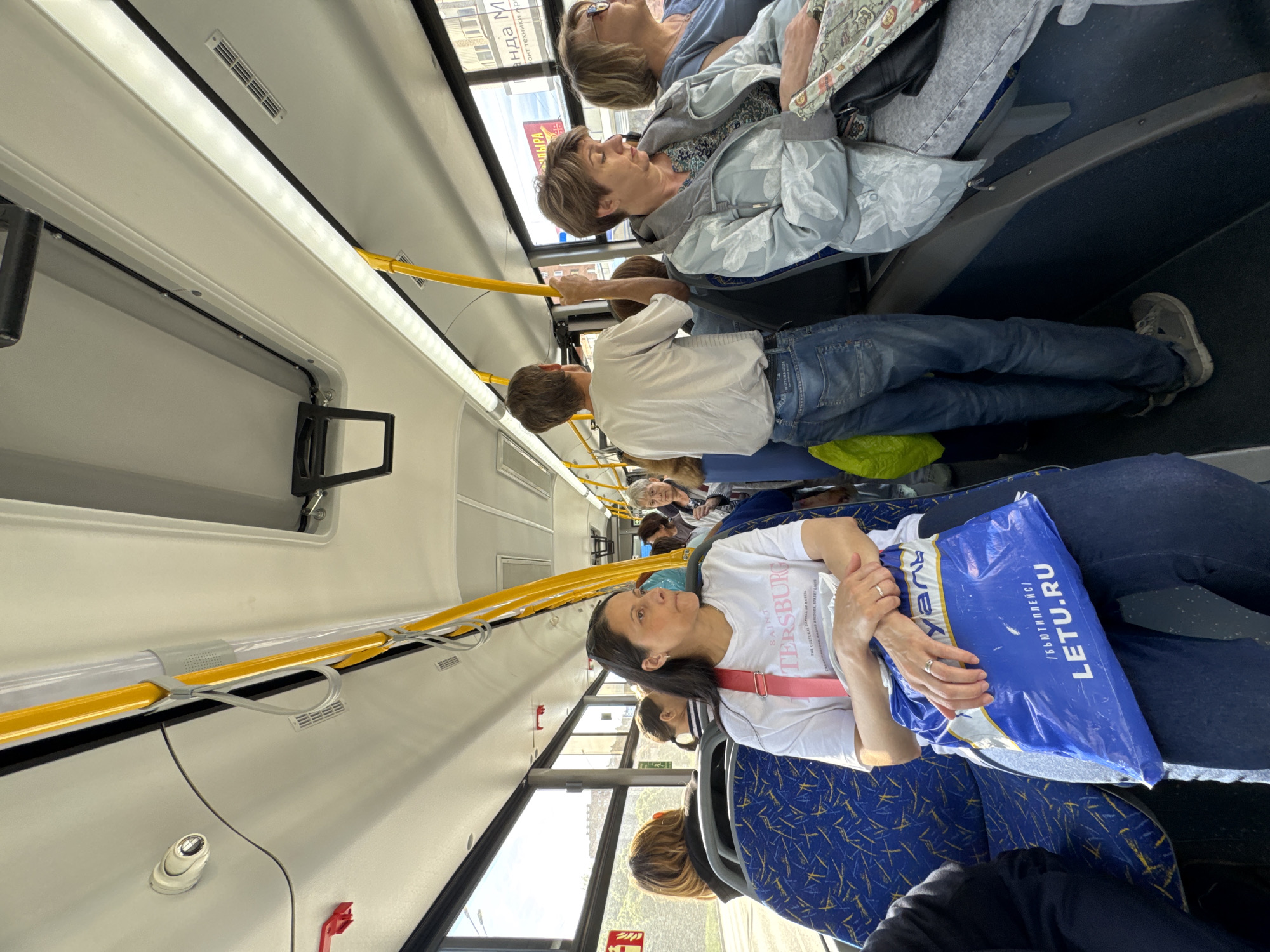 The image captures the interior of a bus, likely during daytime due to the daylight streaming in through the windows. Multiple passengers are seated or standing along the aisle. In the foreground, a woman with dark hair is seated, wearing a white t-shirt with the word "ST. PETERSBURG" written on it, as well as a blue plastic shopping bag from "LETU.RU". Further back, another passenger, a man in a white shirt and jeans, is holding onto a yellow handrail. Other passengers include two women sitting next to each other. The bus interior is a combination of white walls and a blue and yellow patterned seating. The presence of Russian writing on the shopping bag suggests that the bus is likely located in Russia. The windows show a cityscape.