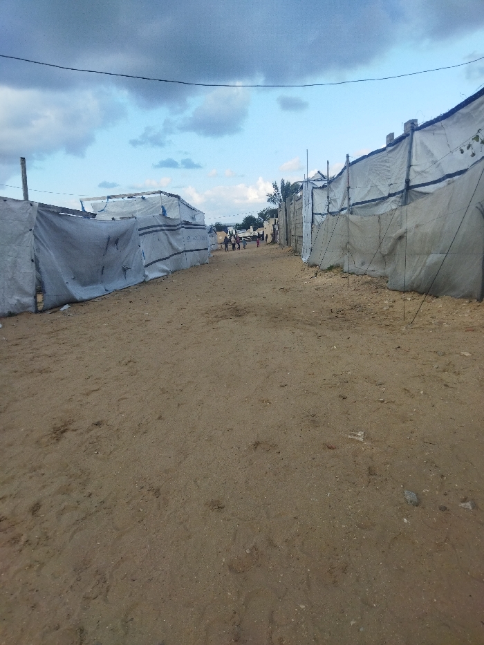 A dirt road leads between makeshift dwellings covered in white material. Some people are walking further down the road. The structures are supported by poles and ropes, suggesting a temporary or informal settlement. The sky is blue with scattered clouds, hinting at daylight. The ground is sandy and uneven, with small rocks and marks. An overhead power line is visible.