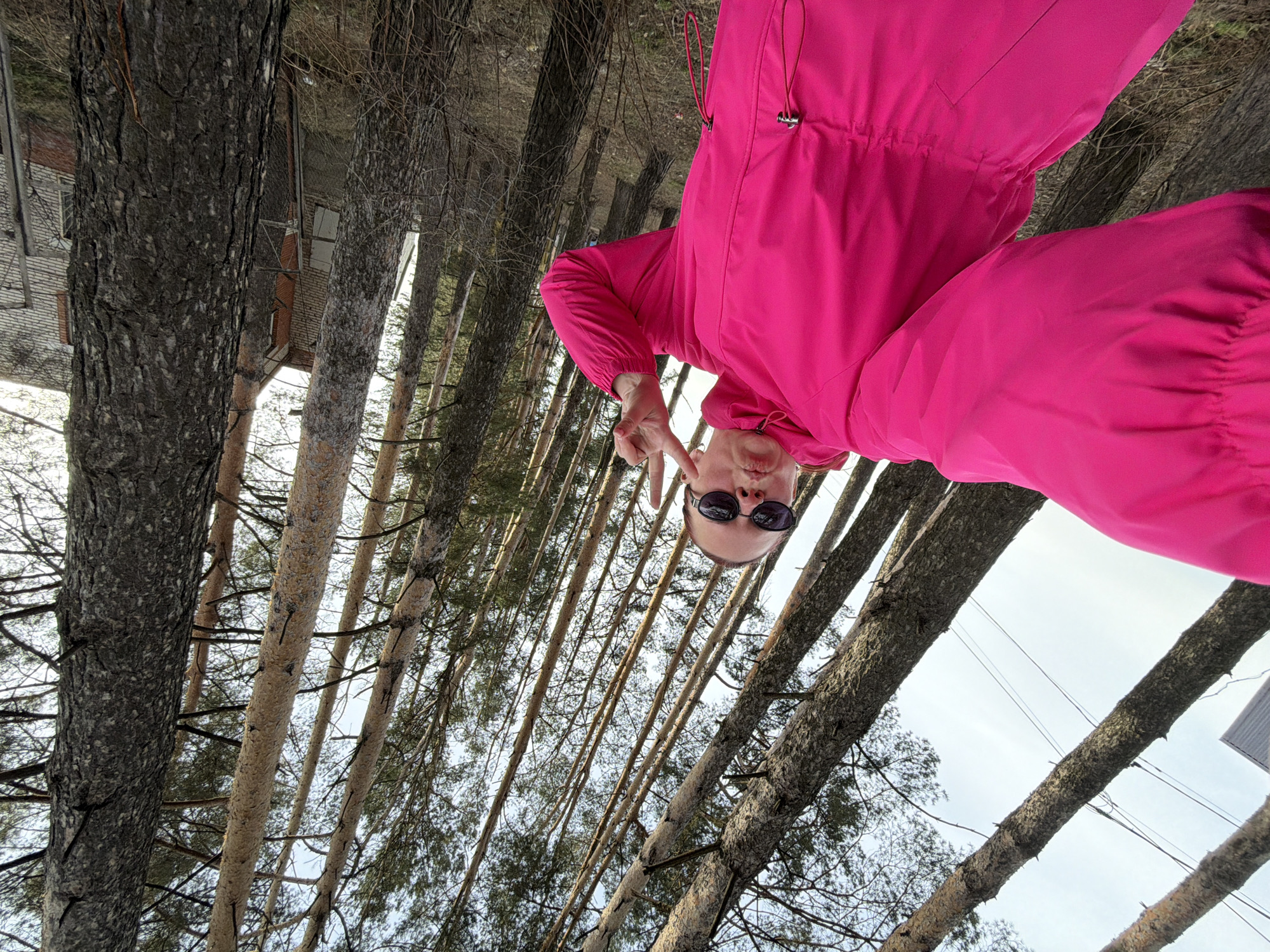 A person wearing sunglasses and a bright pink jacket is giving a peace sign in a forested area. The person is positioned so the trees in the background appear to be growing upwards, suggesting the photo was taken with the camera tilted or inverted. The presence of tall, slender trees indicates a coniferous forest, or at least a wooded area with similar tree types. The location of Chaikovsky, Russia is unknown.