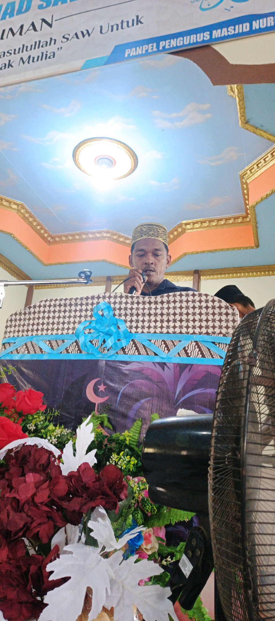 In Simbur Naik, Indonesia, a man wearing a kufi cap stands behind a patterned podium, speaking into a microphone. The podium is draped with fabric featuring a crescent moon and star motif, and decorated with blue ribbon and artificial flowers, including red roses and white leaves. Behind the man, a banner with text in Indonesian is visible, along with a painted ceiling depicting a sky with clouds and a light fixture. To the right, a black oscillating fan is positioned in front of the podium. Another person is partially visible in the background, wearing a black cap. The overall setting appears to be an indoor event or ceremony.