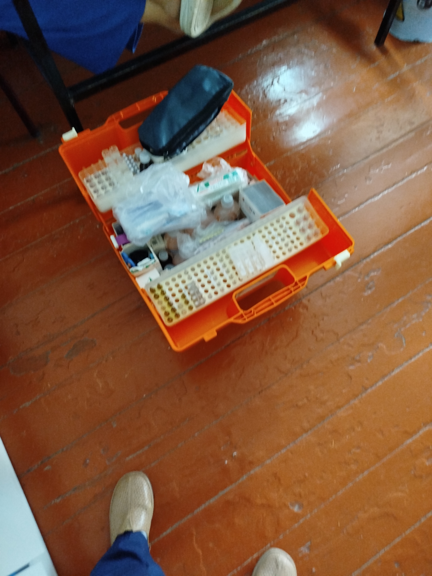 An overhead, slightly blurry view shows an open orange plastic medical kit on a wooden floor. The kit contains various medical supplies, including vials, syringes, and what appears to be a small black pouch. The floorboards are old and show signs of wear. In the bottom left corner, a person's leg in blue pants and a beige shoe is visible, suggesting they are standing near the kit. The scene is indoors, likely in a room with natural light reflecting off the floor. The location is Orozbekov, Kyrgyzstan.