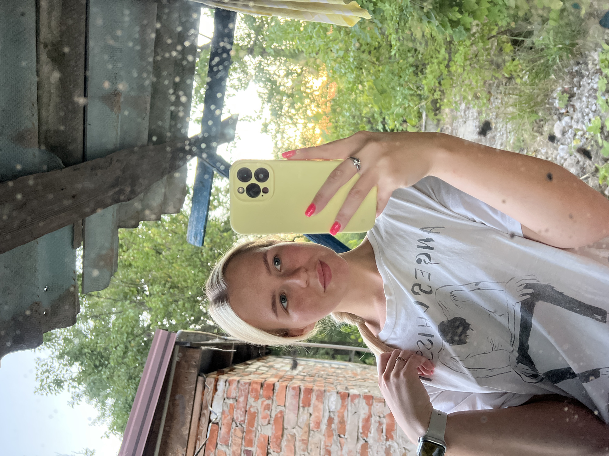 A young Caucasian woman with blonde hair in a braid is taking a mirror selfie. She is wearing a white t-shirt with a graphic print and a ring on her right hand. She is holding a yellow iPhone in her right hand. The background appears to be an outdoor setting with greenery and a brick wall, possibly in a rural or suburban area. The lighting suggests it is either early morning or late afternoon.