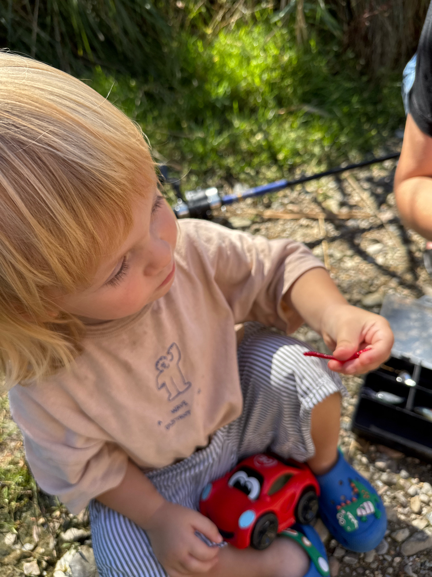The image shows a close-up view of a young child with blonde hair sitting outdoors on a gravelly surface with green foliage in the background. The child is wearing a light-colored t-shirt with a blue graphic of a bear and striped pants. The child's right hand is holding a small red object, possibly bait, and is near a small black tackle box. A red toy car is visible on the child's lap, and the child is wearing blue Crocs with dinosaur designs. In the background, a fishing rod is partially visible. The overall scene suggests an outdoor activity, possibly fishing, taking place in a natural or semi-natural environment. The location is described as Penzano, Italy.