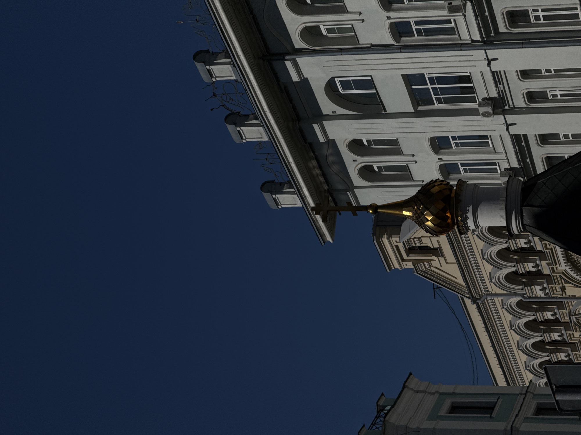The image shows a building in Moscow, Russia, viewed from below. The top of an onion dome, presumably from a church, is visible. The architecture is classical, with arched windows and elaborate detailing. The sky is a deep blue.
