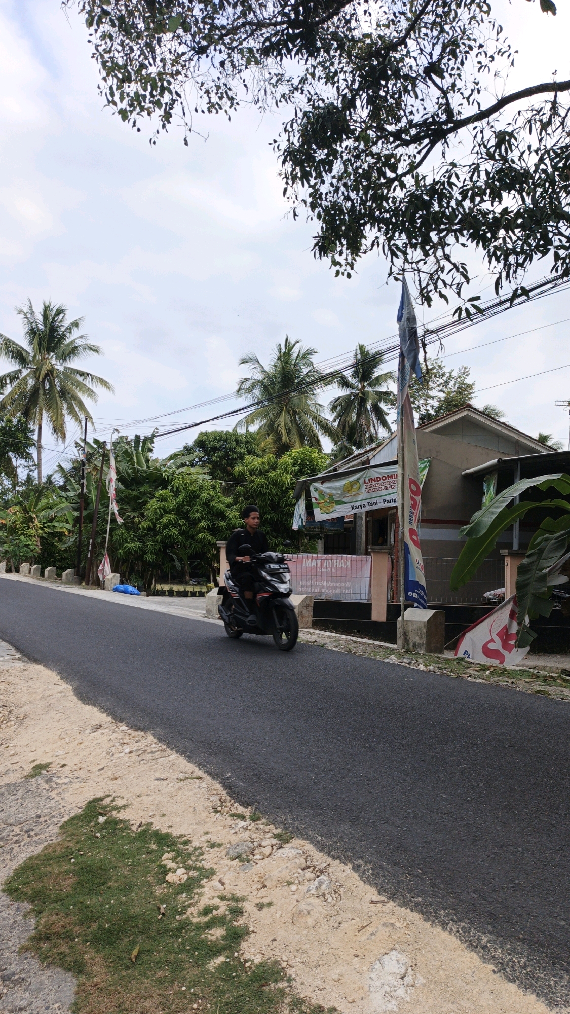 A man rides a black scooter on an asphalt road in Cintaratu, Indonesia. The road is lined with sand and grass on the left, and on the right, there is a building with a covered porch and several banners. Tropical trees and palm trees are visible in the background, and the sky is partly cloudy. The man is wearing a black shirt and shorts and appears to be looking ahead. A flag is planted on a pole to the right of the road in front of the building.