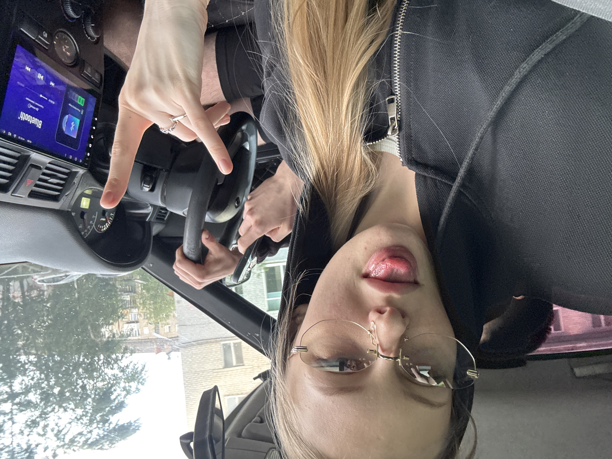 A person is positioned inside a vehicle, possibly the driver's seat, with their face visible and tongue extended. They are wearing glasses and a black jacket with a zipper. Their hair is blonde. One hand is making a hand gesture near the steering wheel. The vehicle's dashboard and infotainment screen are visible, displaying a Bluetooth connection. Through the windshield, a streetscape is visible with trees and buildings. While it is difficult to identify the location definitively from the visual evidence, the style of buildings suggests a European-style urban area. The location is not in Perm, Russia.