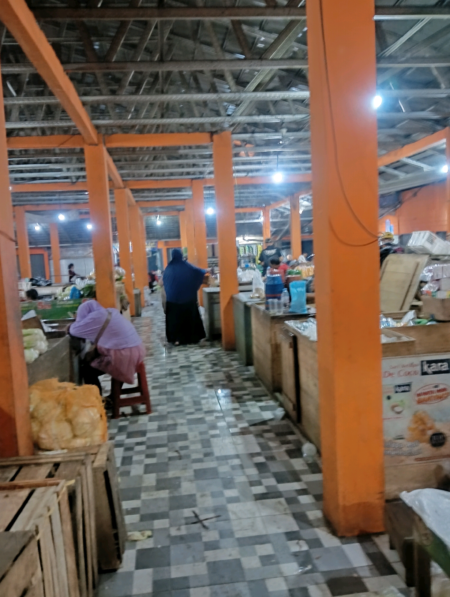 The image depicts the interior of a market hall in Karangjati, Indonesia. The structure features an exposed wooden roof with metal trusses and is supported by prominent orange-painted pillars. The floor is tiled in a grey and white checkerboard pattern, and the walkway is lined with wooden stalls. In the foreground, stacked wooden crates are visible, along with a large, pale yellow object wrapped in plastic. Further into the market, several individuals are present. One person, wearing a purple hijab and pink garment, is seated on a red stool, appearing to rest their head on a table. Another individual in a blue hijab and black skirt stands near a stall. Other people are seen in the background, some attending to stalls where goods are displayed. The lighting appears to be artificial, with bright lights illuminating the space. The overall scene suggests a bustling marketplace environment.