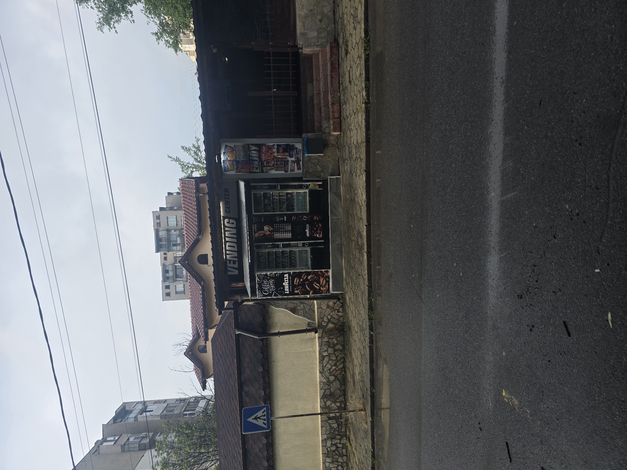 The image captures an outdoor street scene in Radnevo, Bulgaria, depicting a "VENDING CENTER" building positioned alongside an asphalt road and a paved sidewalk. The scene is set during the daytime under a clear, bright sky, suggesting fair weather.
The primary subject is a small, single-story structure housing multiple vending machines, bearing a prominent "VENDING CENTER" sign on its facade. The building has a dark tiled roof and is constructed with a mix of stone cladding and light-colored plaster. Two main vending machines are visible: one for snacks and drinks, displaying the brand "ENJOY" and various packaged goods, and another for coffee and hot beverages, branded "Lavazza," featuring images of coffee beans and the text "Coffee & Drinks." A small blue plastic bin is located next to the snack machine. A dark metal drainpipe is visible on the side of the vending center building.
To the left of the vending center, a blue pedestrian crossing sign, featuring a white triangle with a walking figure, stands on a pole. The asphalt road in the foreground shows a faint white line, and a concrete curb separates it from the paved sidewalk. In the background, beyond the immediate structures, several multi-story residential buildings with tiled roofs and visible windows can be seen under the clear sky, interspersed with some green tree foliage. Multiple black power lines crisscross the upper part of the frame.
