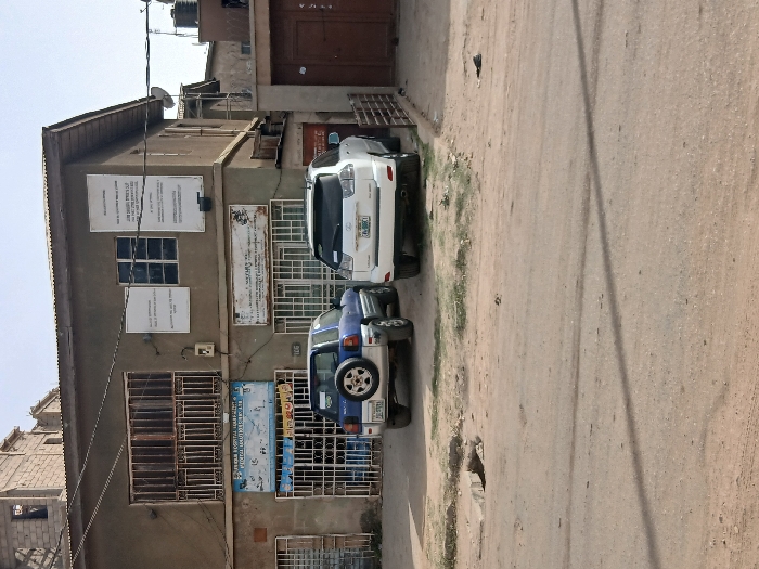 The scene depicts a street view in Mgbidi, Nigeria, featuring a two-story building under a clear sky. The building, constructed with light brown/grey walls and topped with a corrugated metal roof, appears weathered. The ground floor houses two commercial establishments, both secured by metal grilles over their windows and entrances. The left establishment displays a blue sign partially reading "EZEAM MEDICAL DIAGNOSTIC & PHYSIOTHERAPY," with the address "106" visible below. The right establishment has a white sign partially visible, possibly indicating "NATURAL HEALTH." An electrical meter box is affixed to the wall between these two units. The upper floor features windows protected by ornate metal grilles and three rectangular white signs with text. Behind the main building, an unfinished concrete block structure with exposed rebar is visible.

Two sport utility vehicles are parked on the unpaved dirt and gravel road in front of the building. On the left is a blue Toyota RAV4, an older model, with a spare tire mounted on its rear hatch; its license plate is partially legible, beginning with "KGE." On the right is a white Lexus RX SUV, with its license plate partially legible, ending with "FBC."

The foreground consists of an uneven, unpaved road surface with scattered dry vegetation. A broken concrete edge or small ditch is visible on the left side of the road. Overhead, power lines, a satellite dish, and a black water storage tank on a stand are visible near the roofline on the right side of the building. No individuals or specific activities are present within the frame.