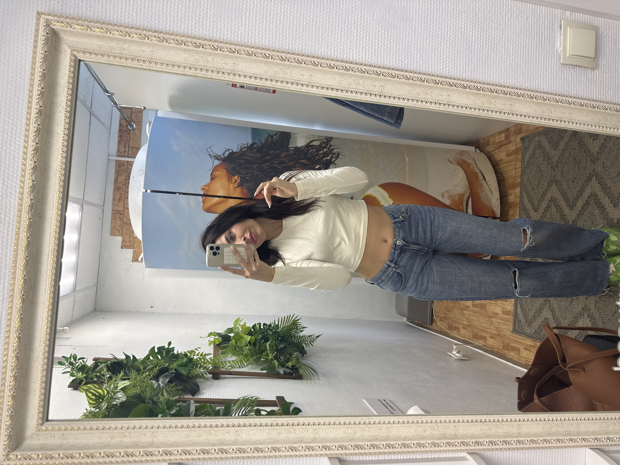 A woman takes a selfie in a mirror in a room in Chaikovsky, Russia. The woman is wearing a white long-sleeve shirt and blue jeans with rips. The room has white walls, plants on a wooden frame, a floor mat, and a brown purse. A poster depicting a woman in swimwear is mounted on the wall behind. A light switch is visible on the upper right.