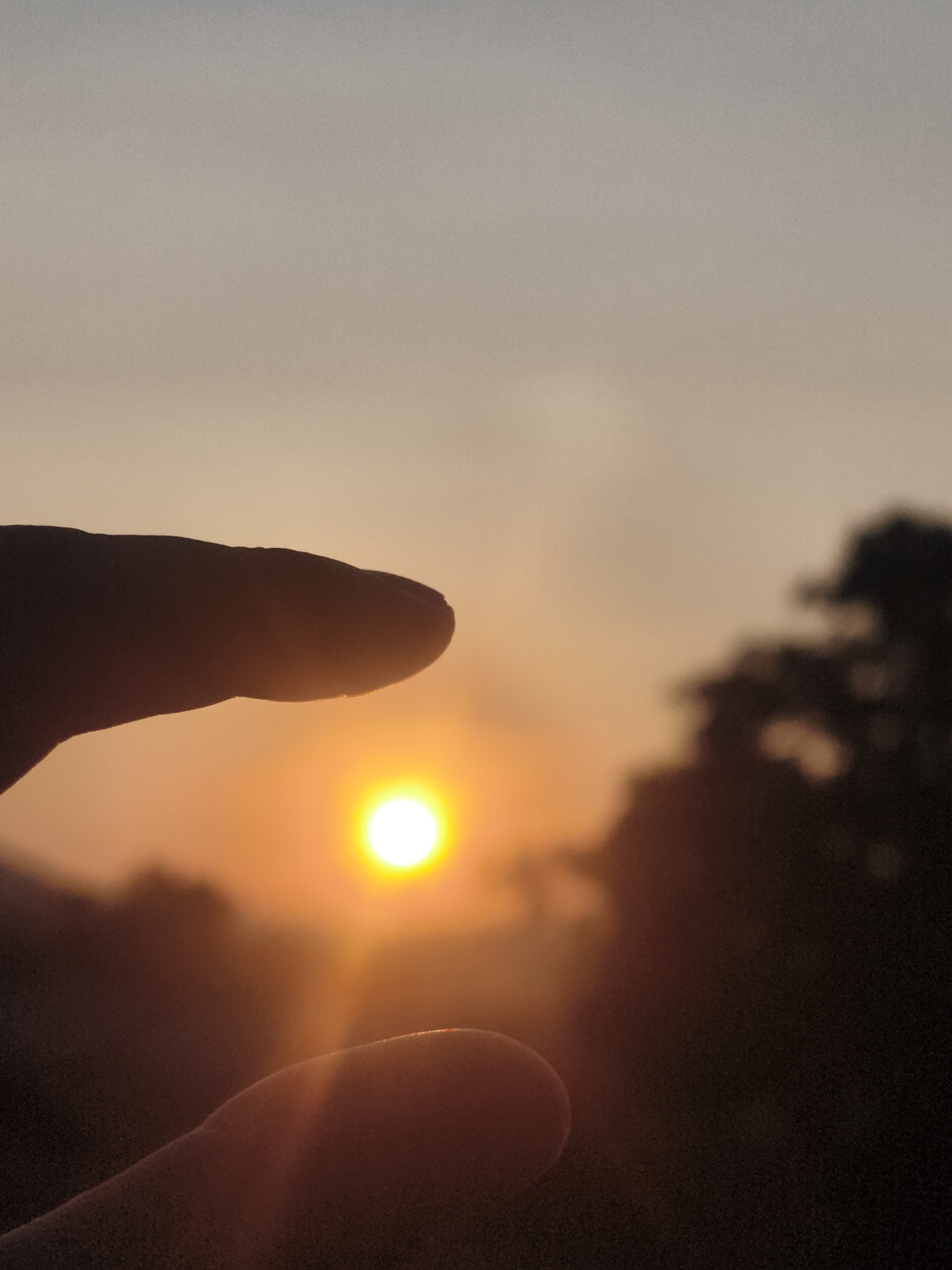 The image shows a hand holding the sun in the city of บ้านบางรักษ์, Thailand. The hand is silhouetted against the bright orange sun. The sky is a gradient of orange and gray, with blurred trees in the background. The sun is a bright, small circle.